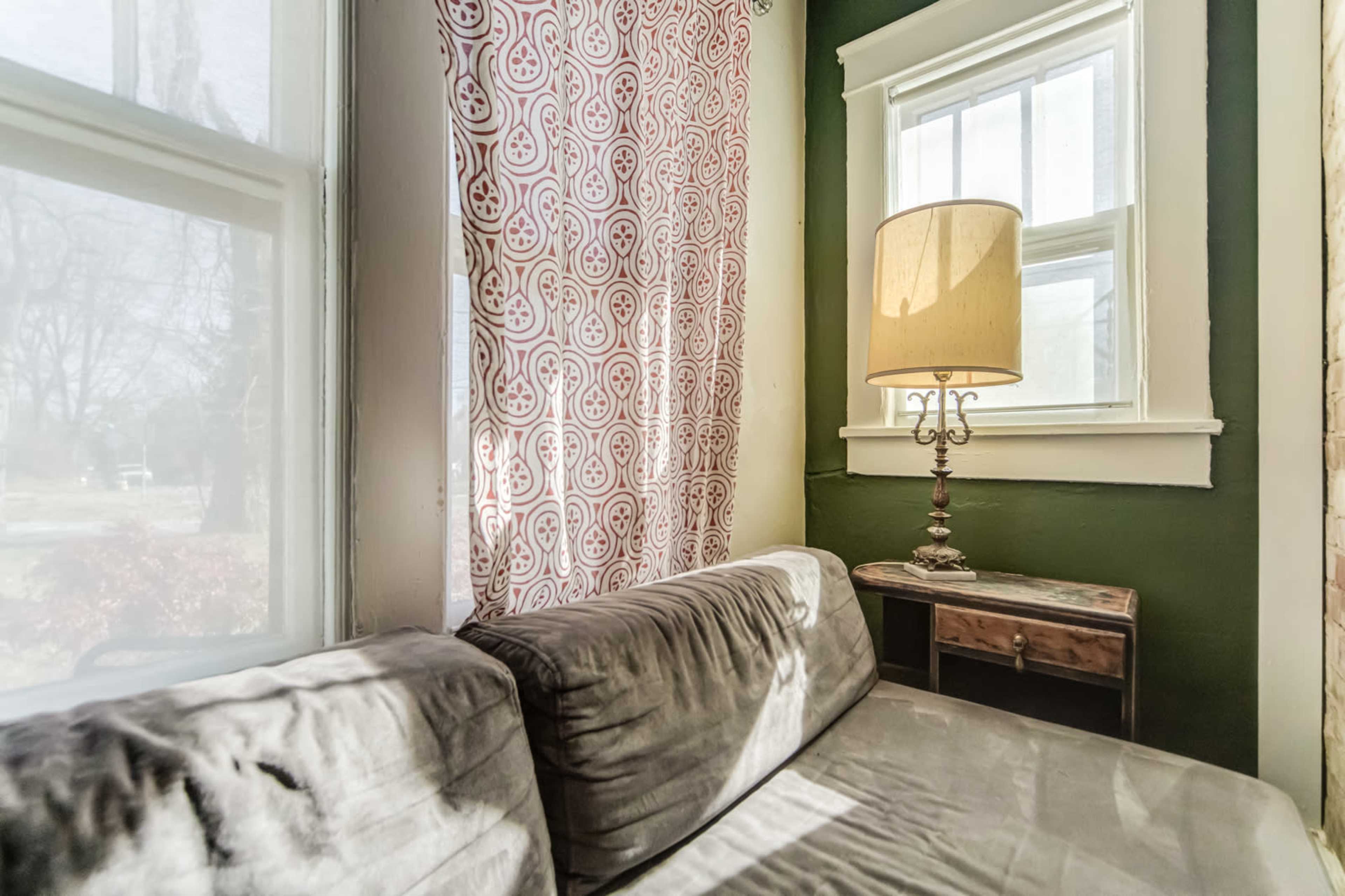 A corner of a room with a gray couch, a standing lamp, a small side table, and patterned curtains next to a window.