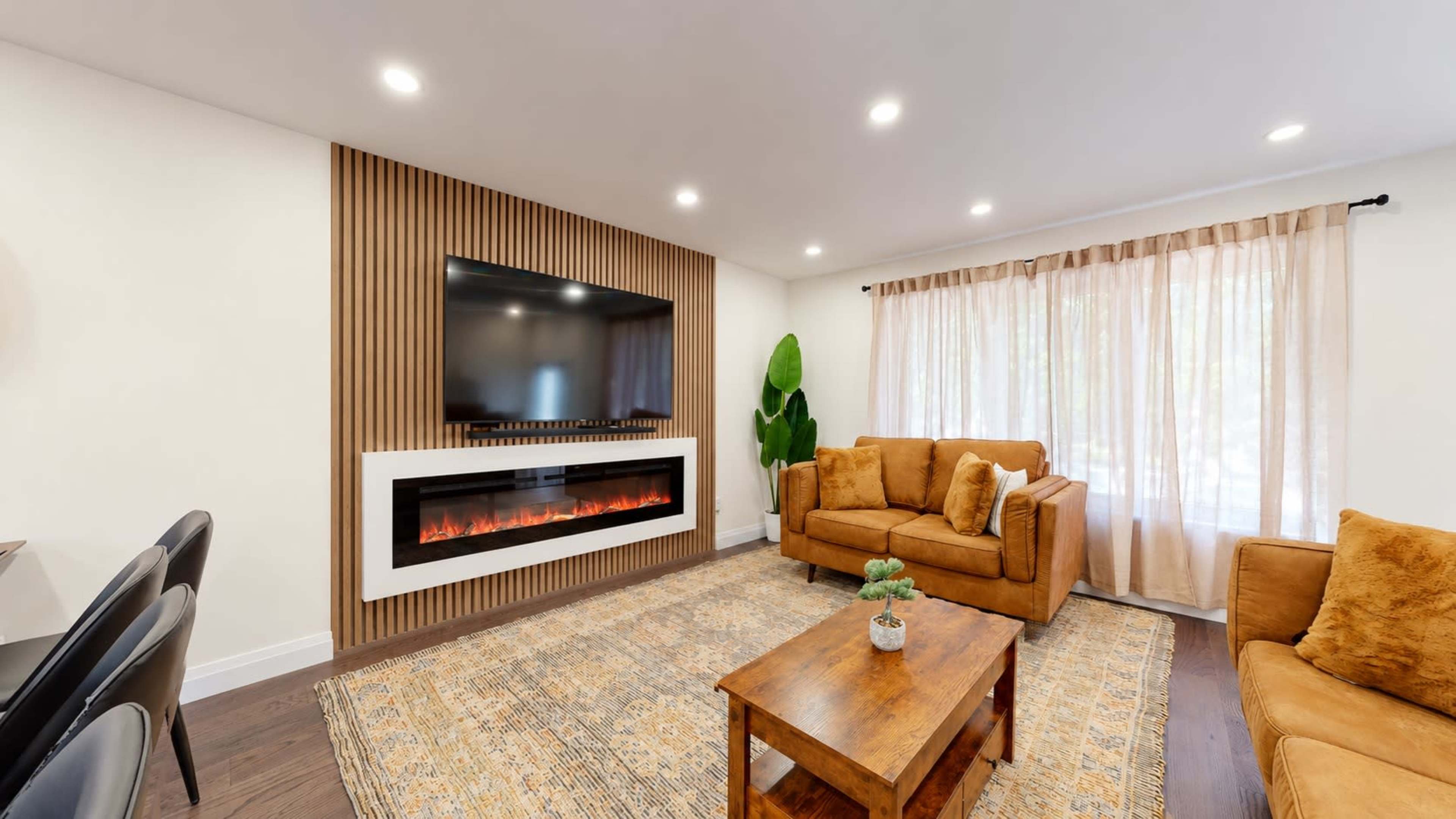 The image shows a modern living room with a mounted flat-screen TV above a sleek white fireplace, complemented by a beige sofa set, a wooden coffee table, and a large window with sheer curtains.