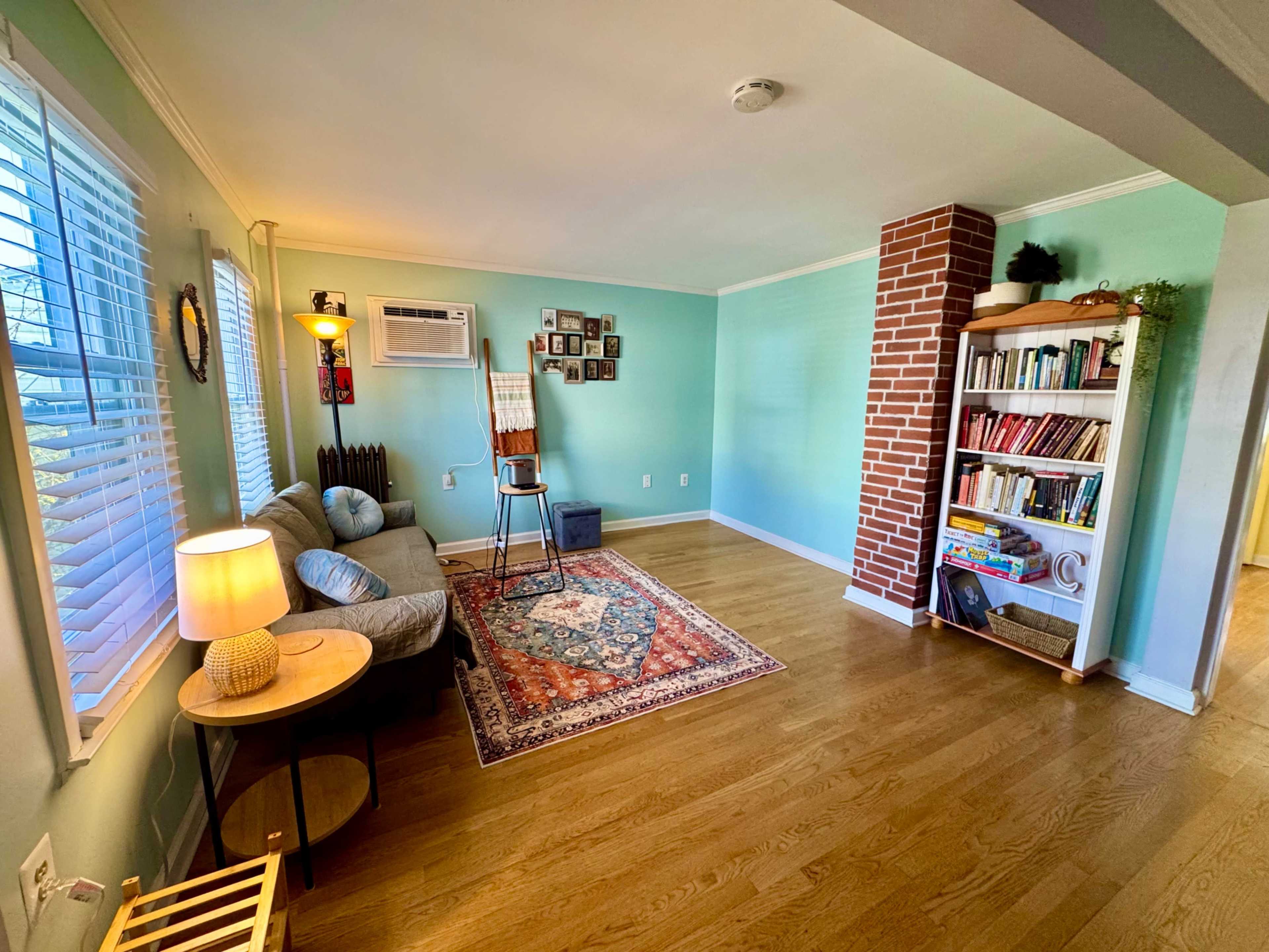 The room features a cozy layout with a sofa, an air conditioning unit, a brick accent wall, and a bookshelf filled with books.