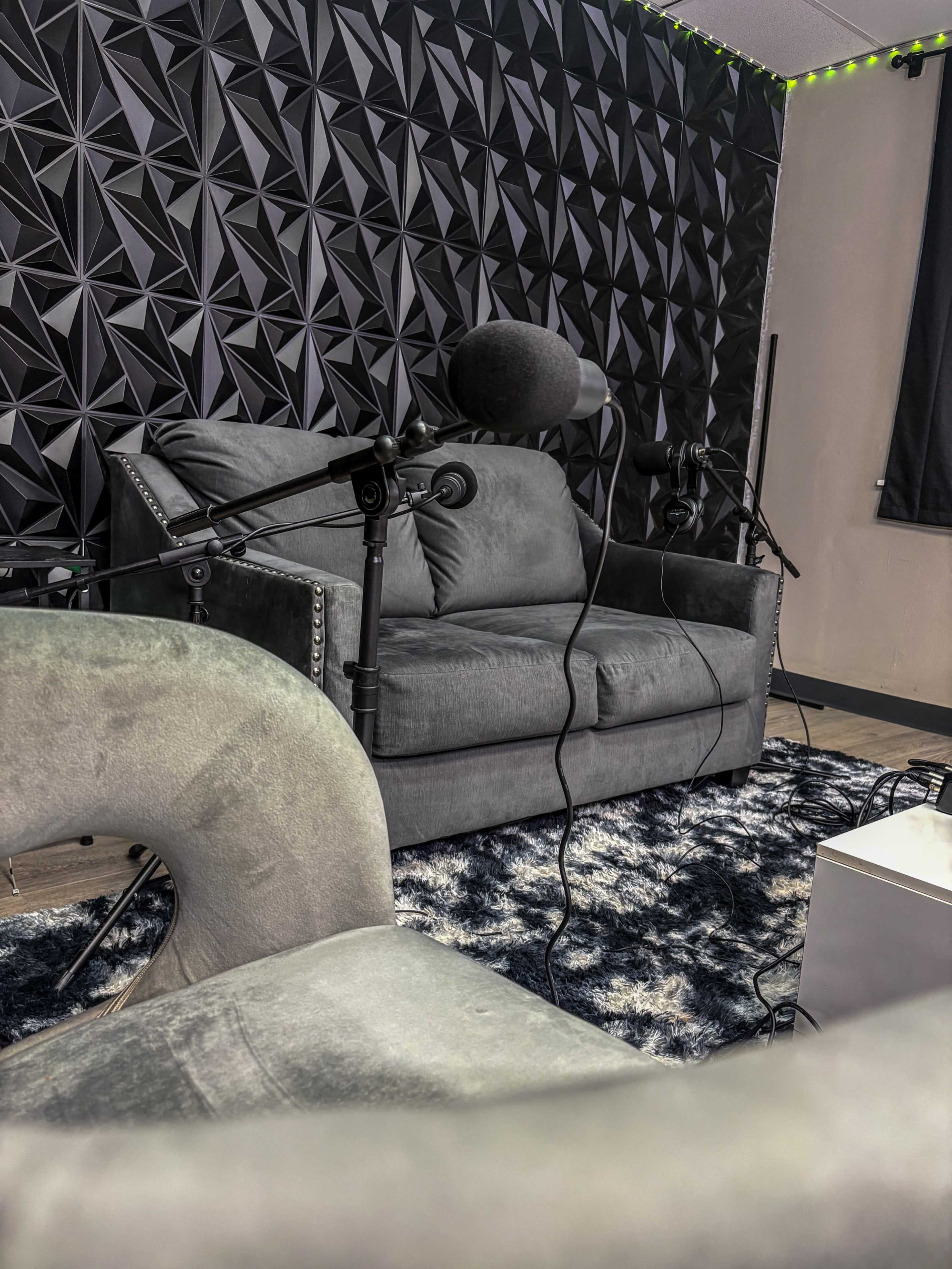 The image shows a recording studio setup with a gray sofa, two microphones on stands, and a plush rug on the floor.