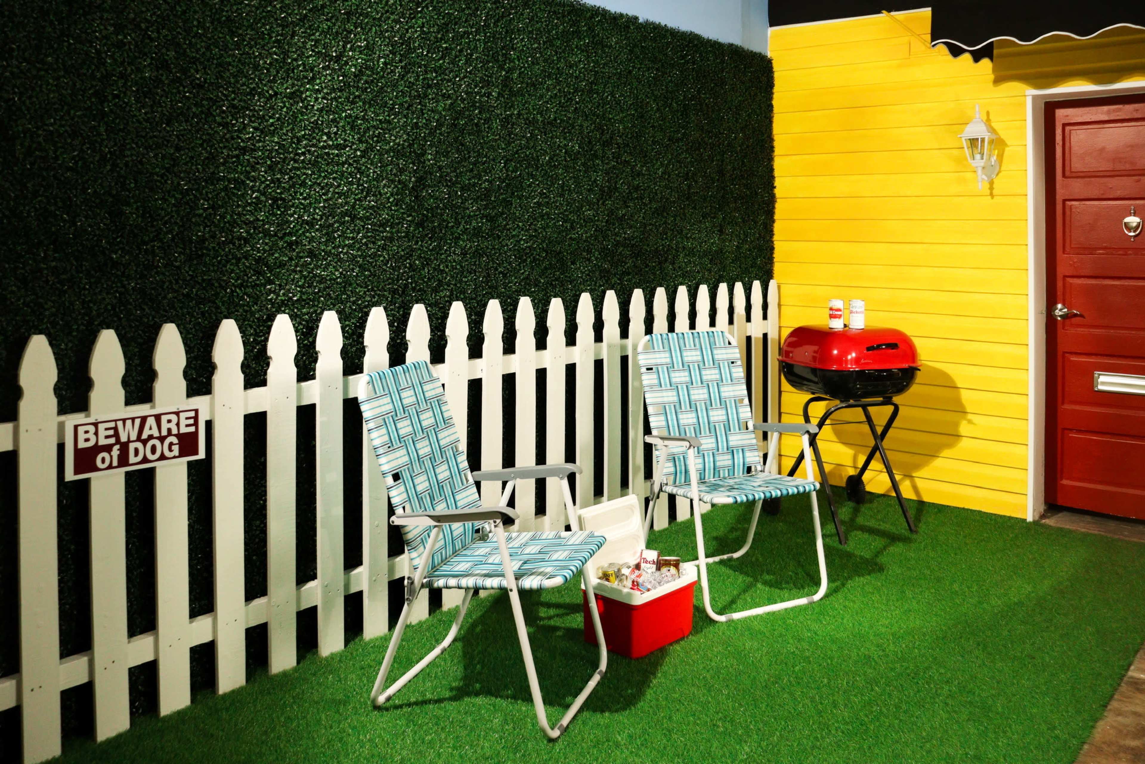 The scene features two folding chairs on green grass in front of a white picket fence, with a yellow wall and a red door in the background, alongside a small grill.