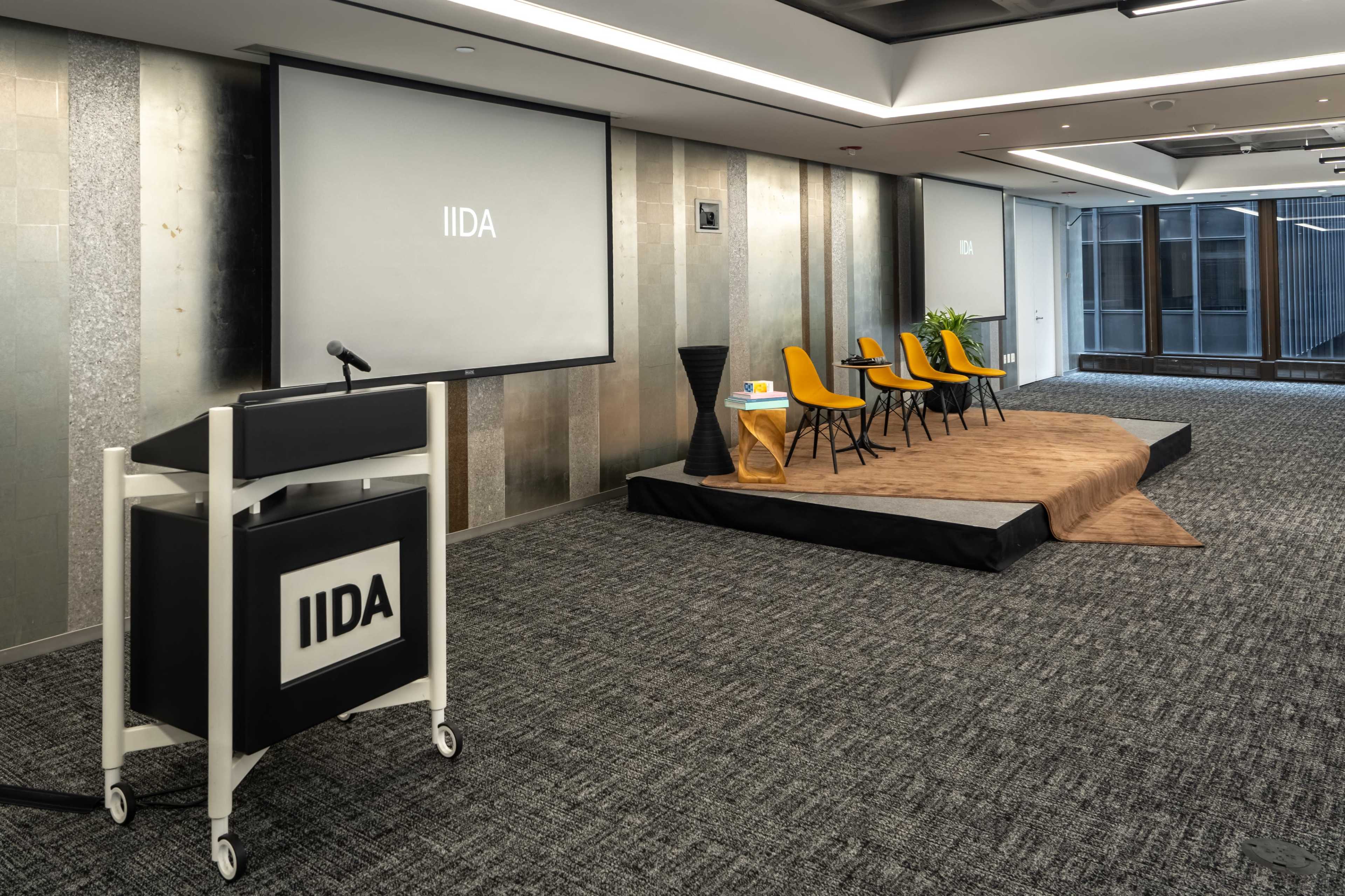 The image shows a modern conference room setup featuring a podium, a large screen displaying the letters "IIDA," and four yellow chairs arranged on a raised platform.