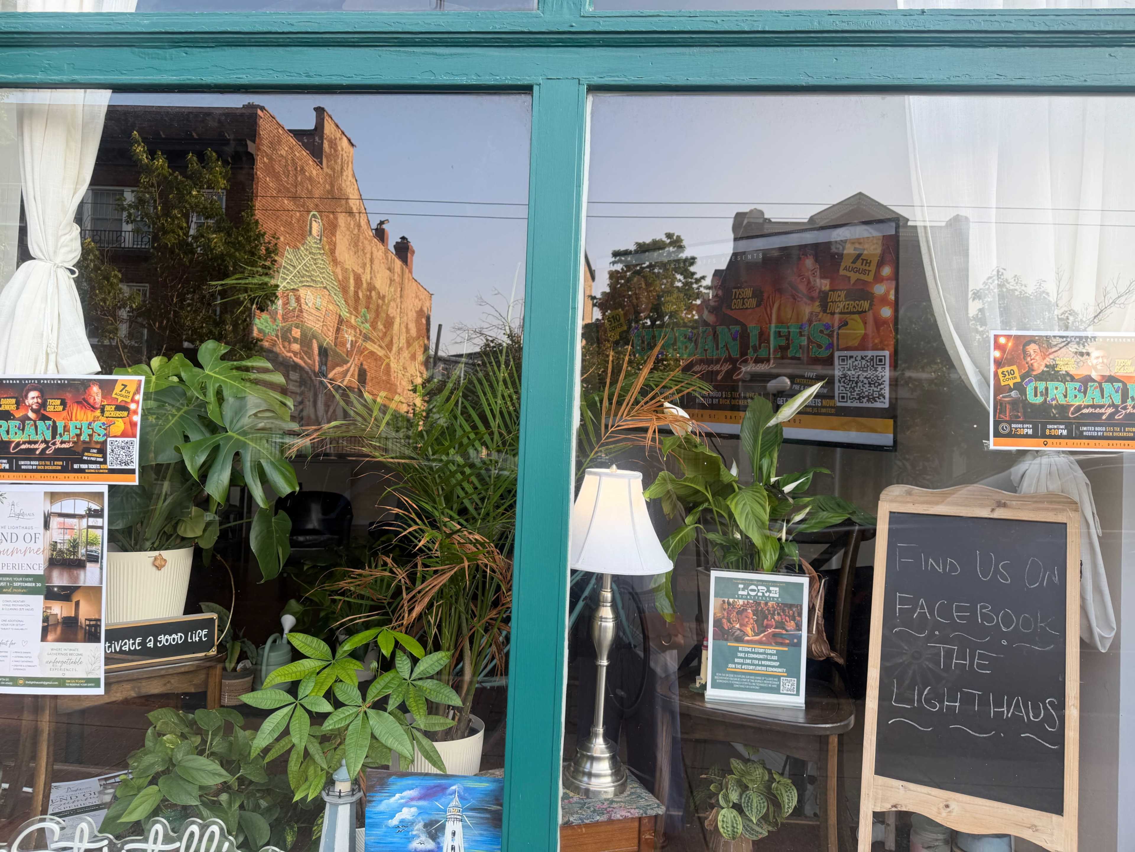 The image shows the exterior of a shop window featuring various indoor plants and promotional signage.