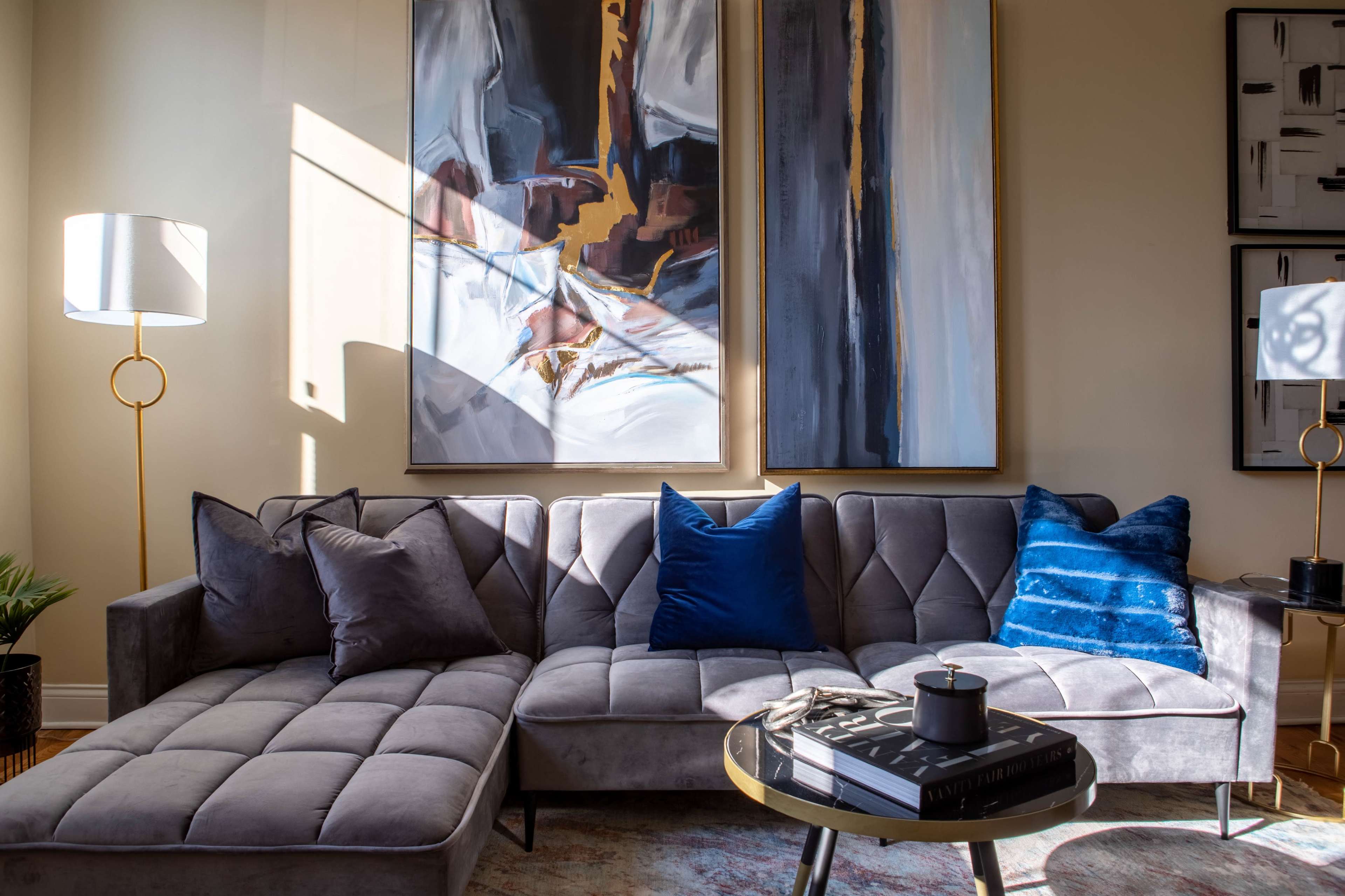 A modern living room features a gray sectional sofa with decorative pillows, a round table with a book, and large abstract paintings on the wall, illuminated by natural light.