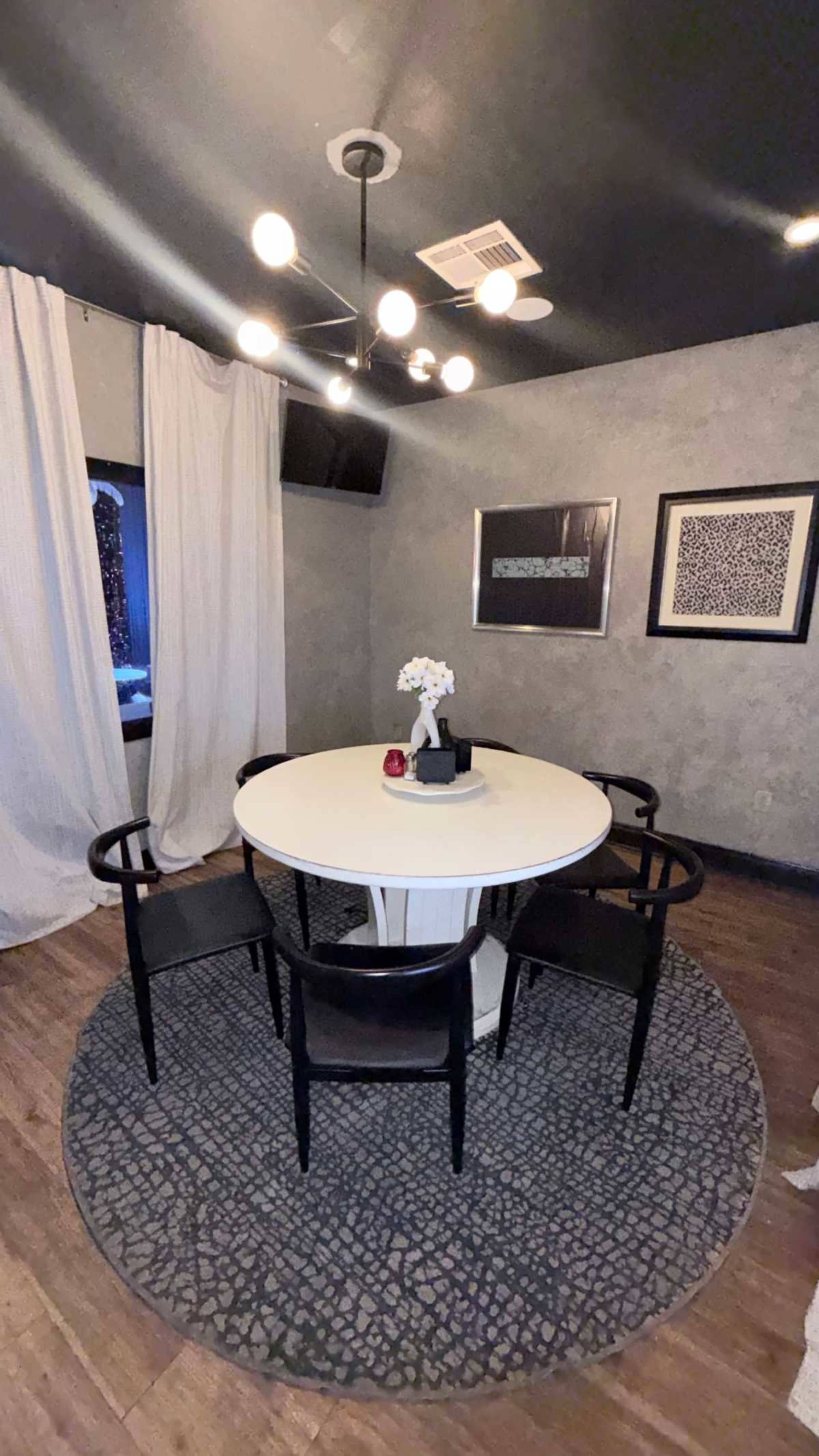 The image shows a modern dining area with a round table, four black chairs, a patterned rug, and abstract wall art, illuminated by a stylish chandelier.