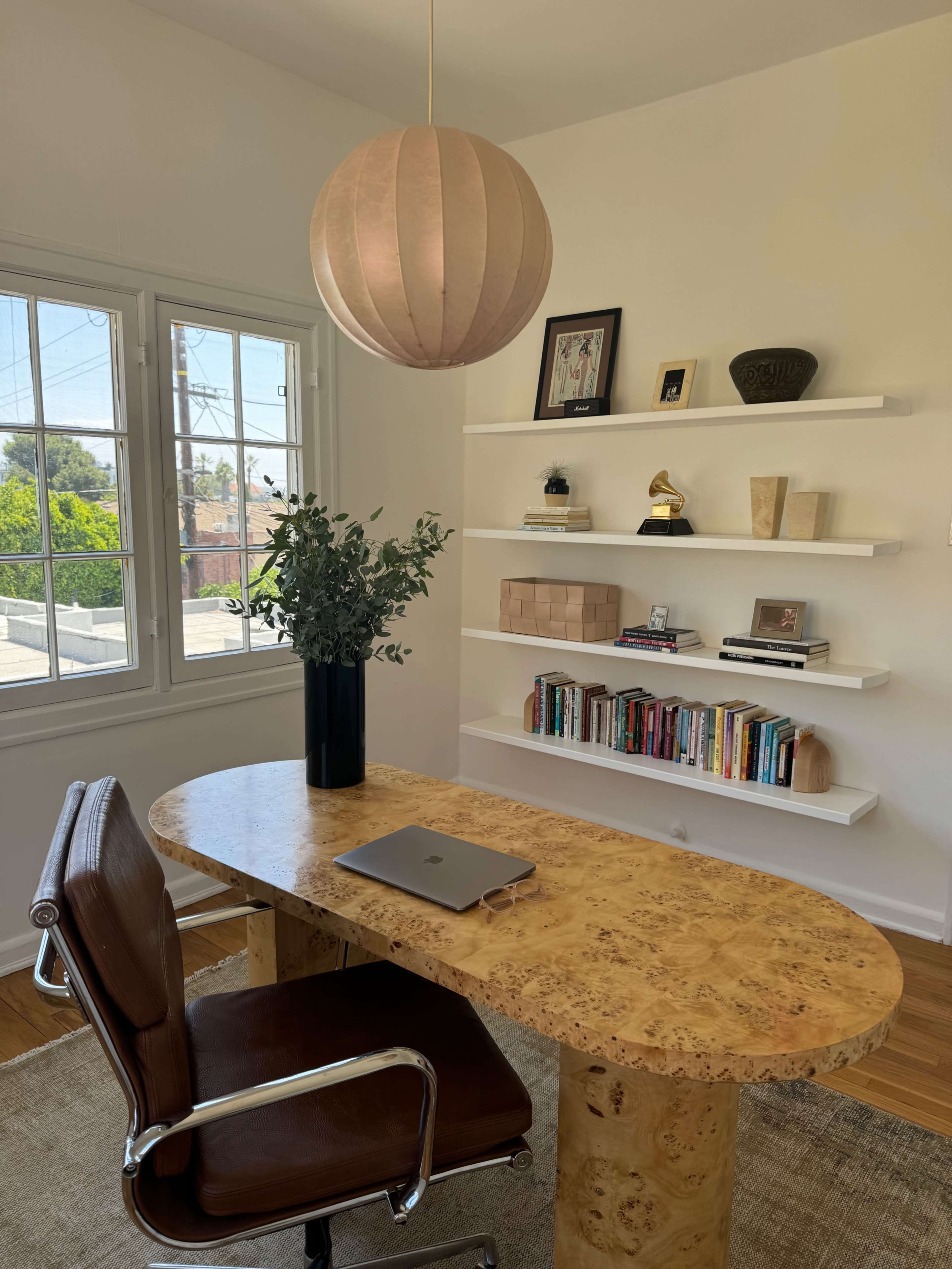 The image shows a modern home office with a round, wooden desk, a laptop on it, and a shelf displaying books and decorative items.