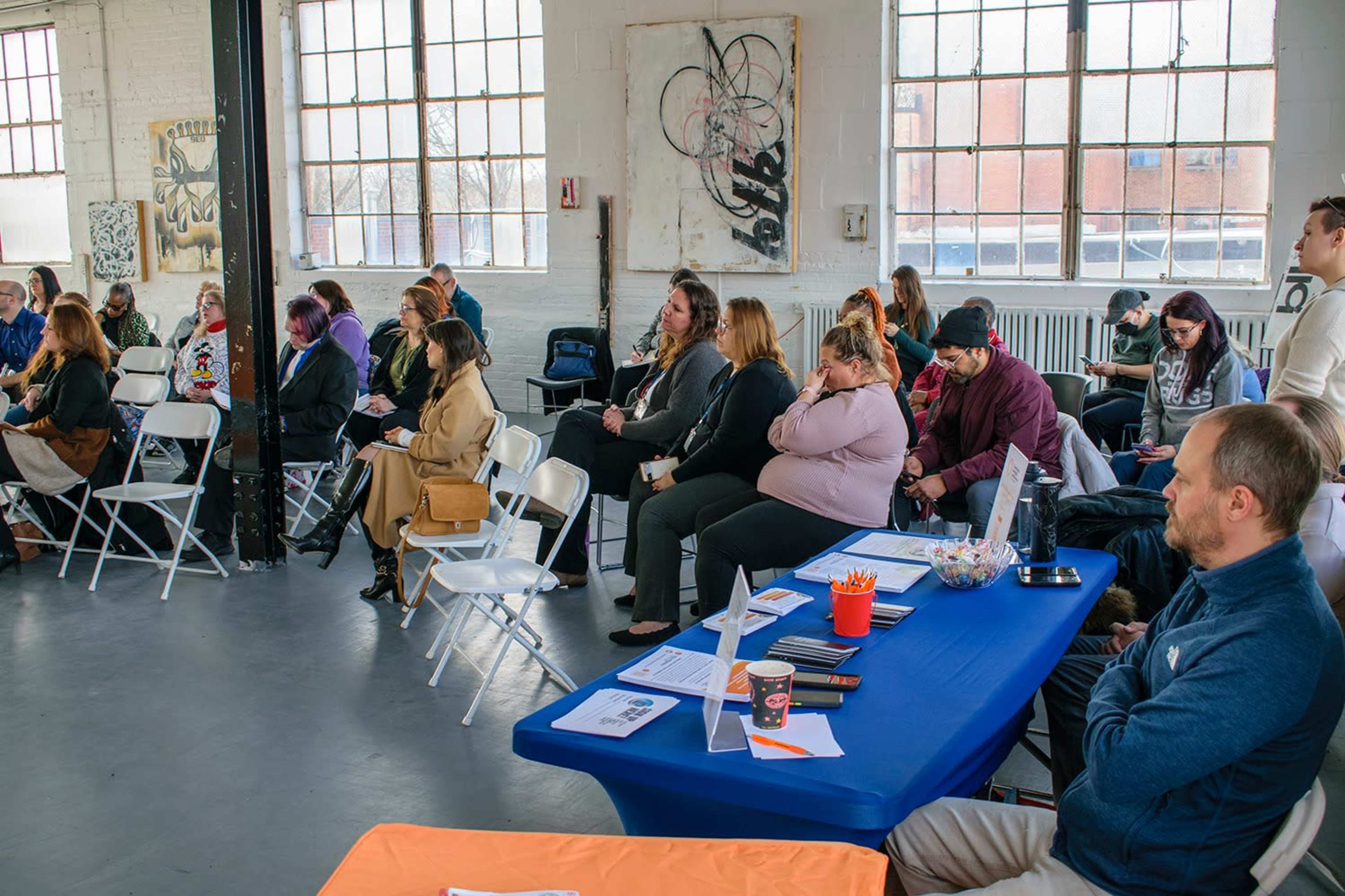 A group of people is seated in an industrial-style room with large windows, participating in an event.