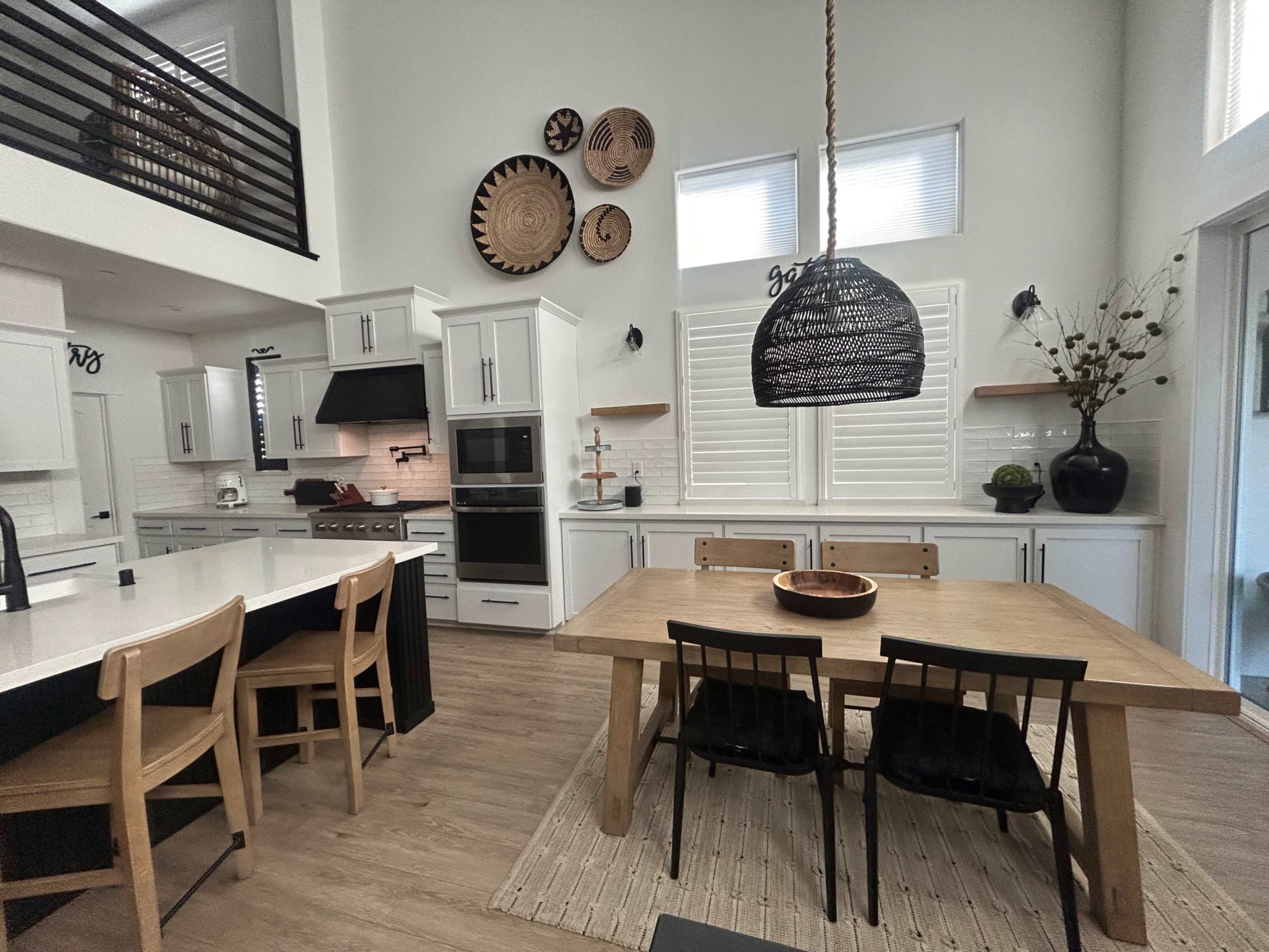 The image shows a modern kitchen and dining area featuring white cabinetry, a wooden dining table, and decorative wall hangings.