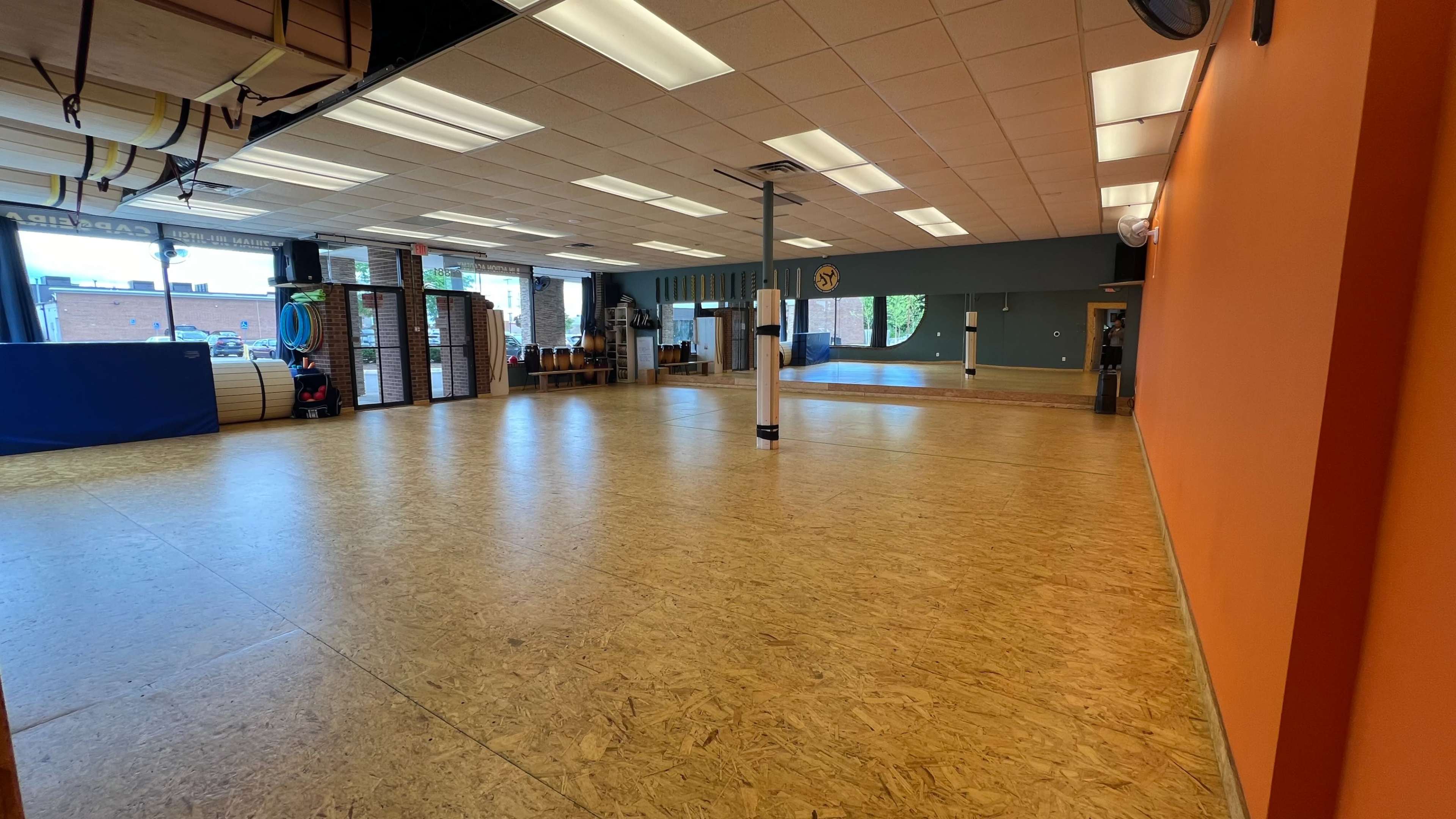 An empty indoor space with wooden flooring and orange walls, featuring various exercise equipment and large windows letting in natural light.