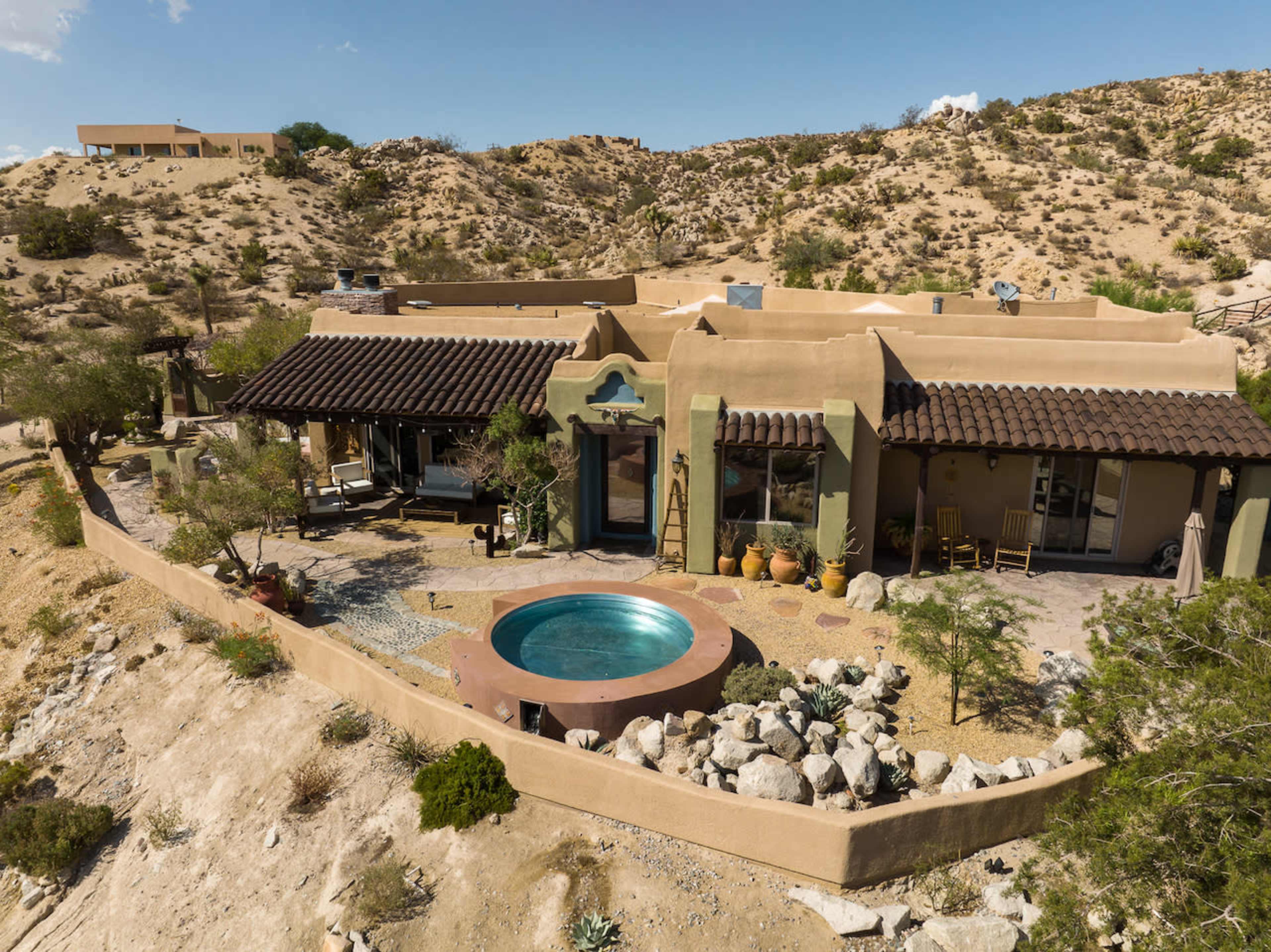 A single-story adobe house with a round hot tub is set in a desert landscape surrounded by sparse vegetation and rocky terrain.
