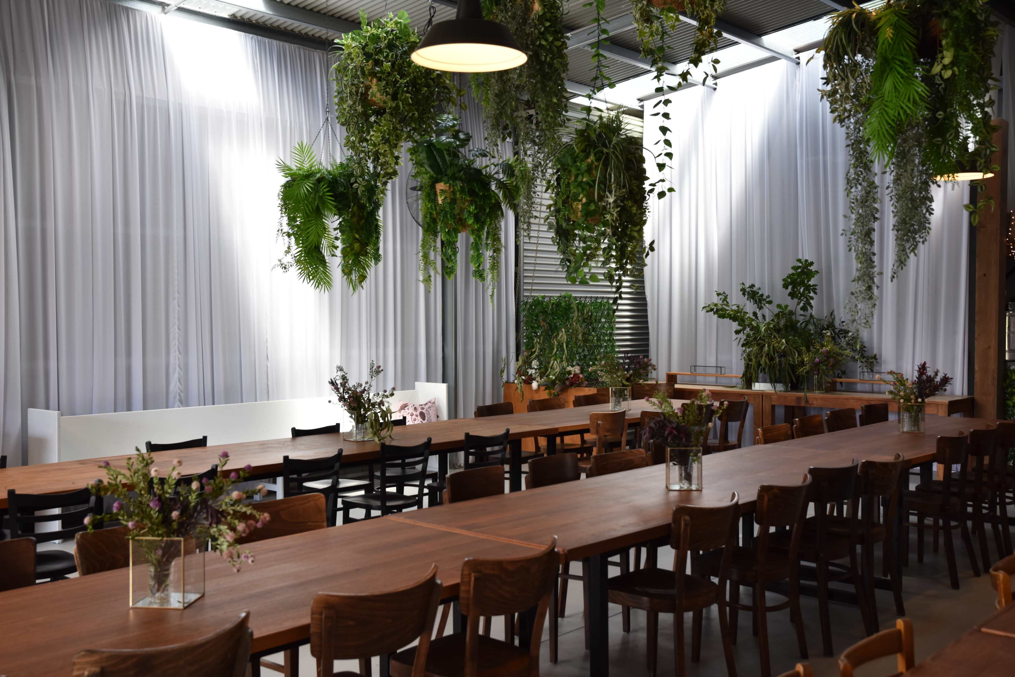 The image shows a spacious indoor dining area with long wooden tables, surrounded by greenery and plants suspended from the ceiling.