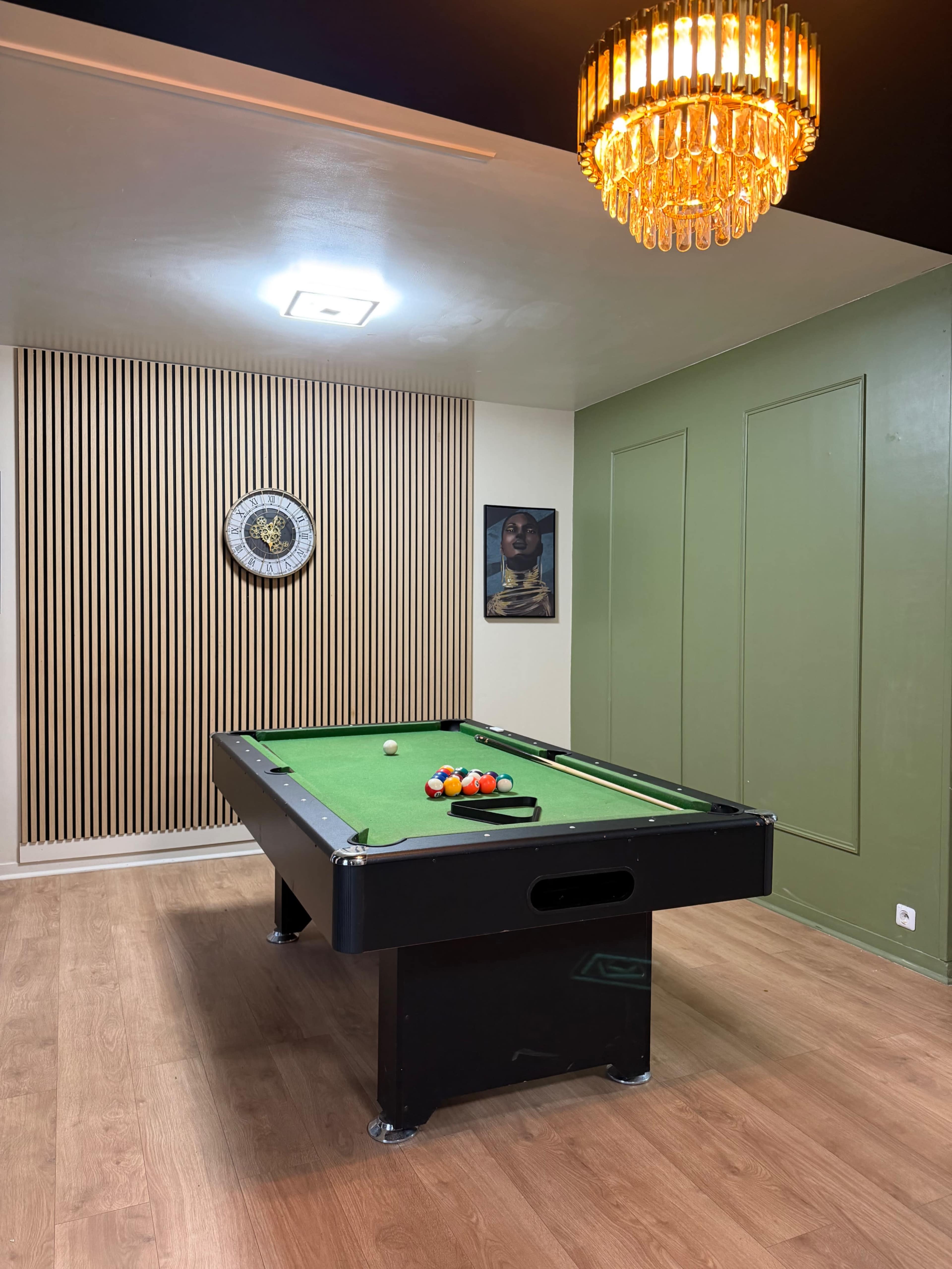 The image shows a game room featuring a green felt pool table, a wall clock, artwork, and a chandelier.