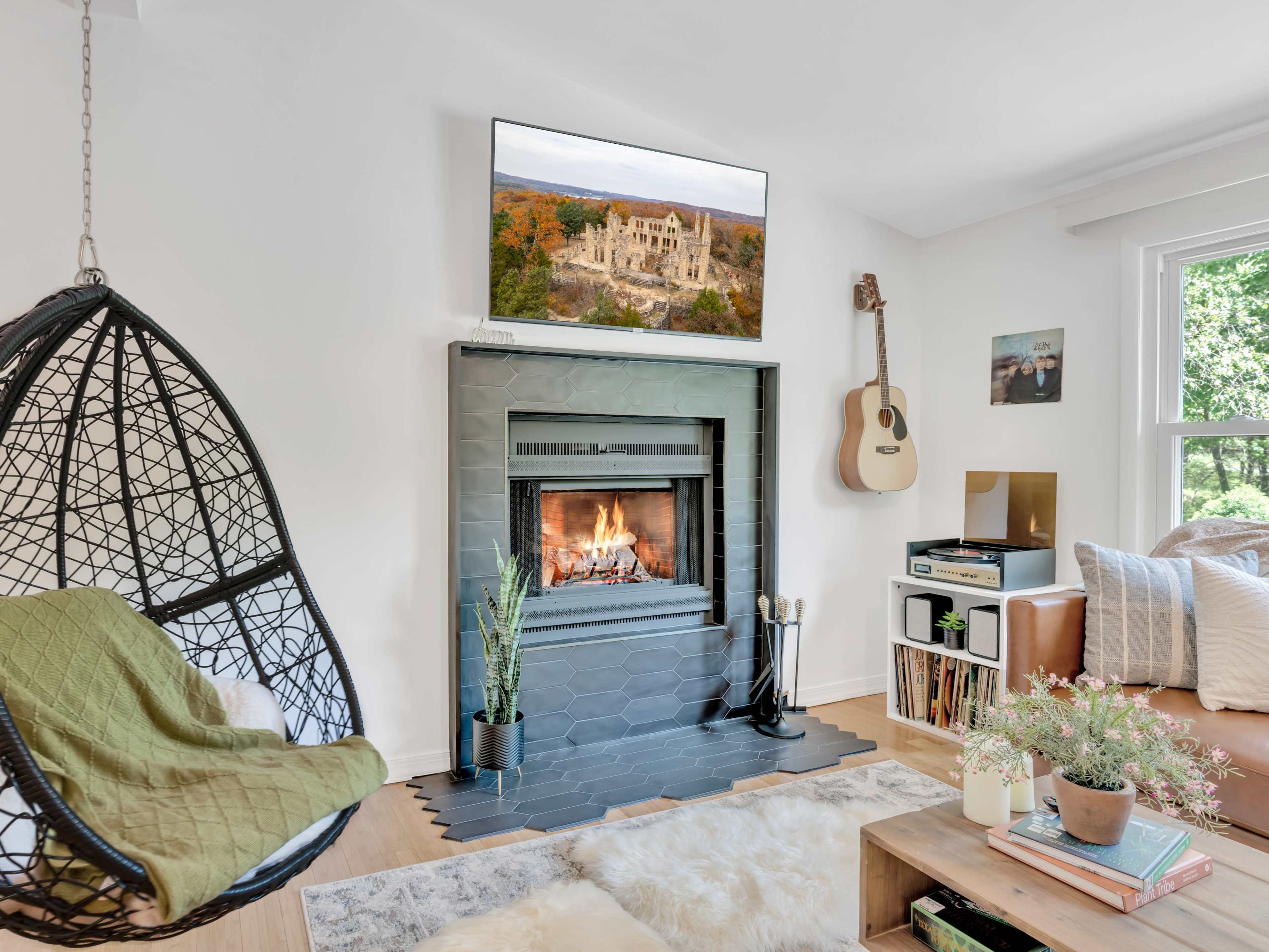 The living room features a hanging chair next to a fireplace with a television mounted above it, accompanied by a guitar, record player, and plants.