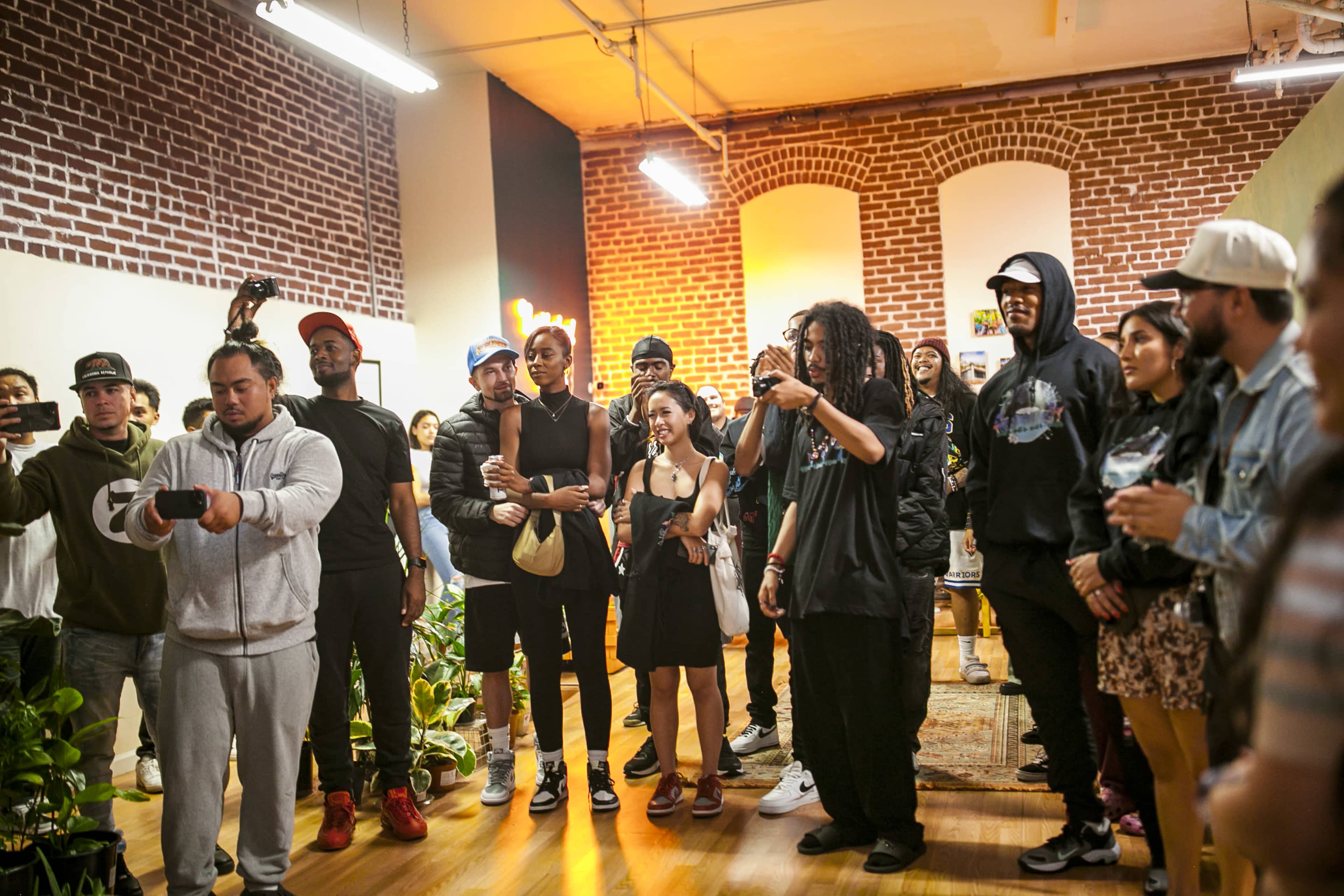 A group of people gathers in a room with exposed brick walls, some holding phones and drinks while watching an event.