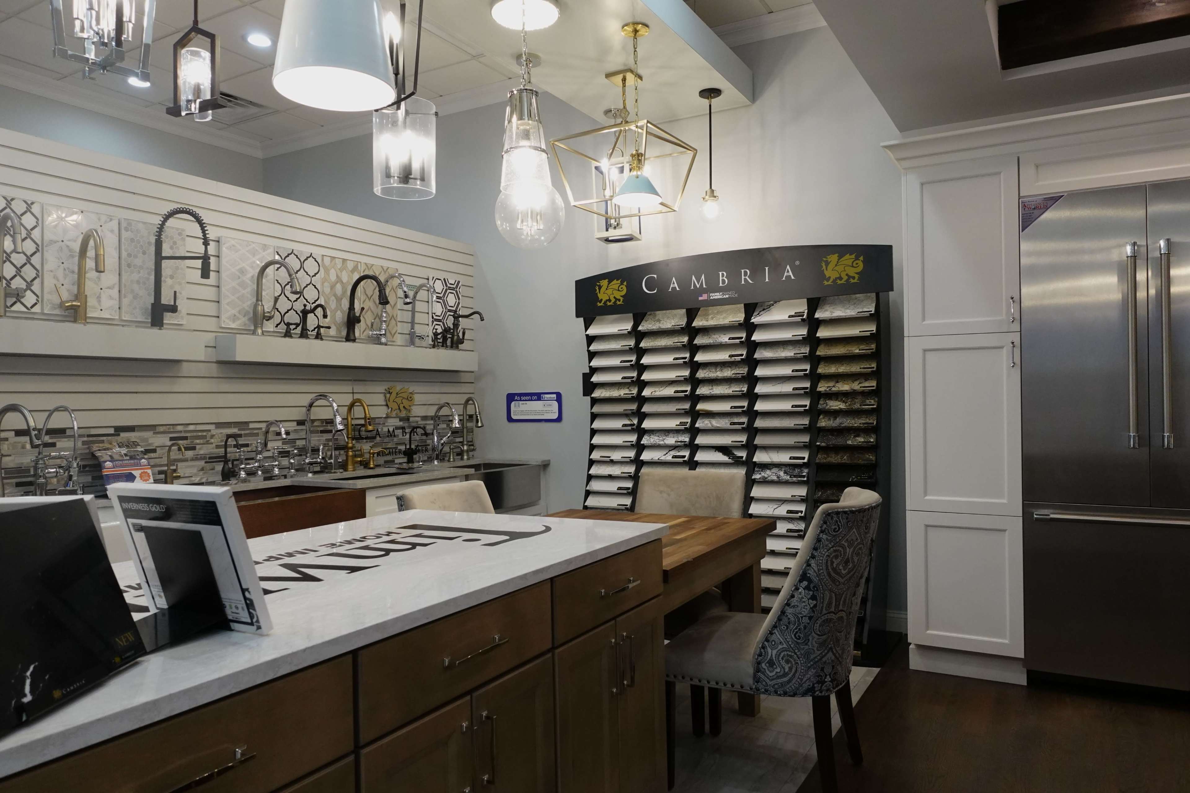 A modern showroom features a display of kitchen faucets and countertop samples arranged on a wall, with a central table and stainless steel refrigerator in the background.