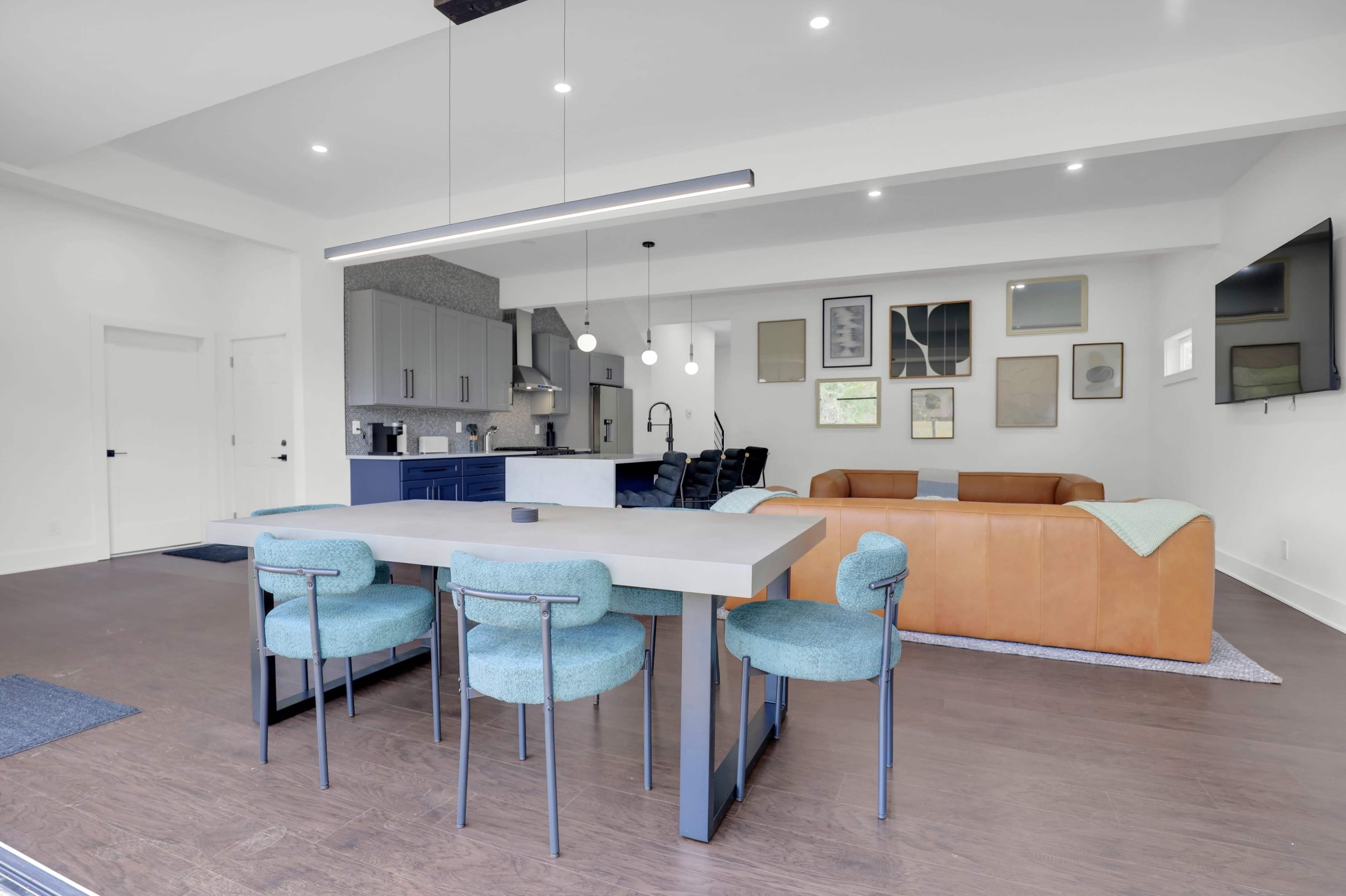 The image shows a modern open-concept kitchen and dining area featuring a rectangular table with teal chairs, a brown leather sofa, and various framed artworks on the walls.