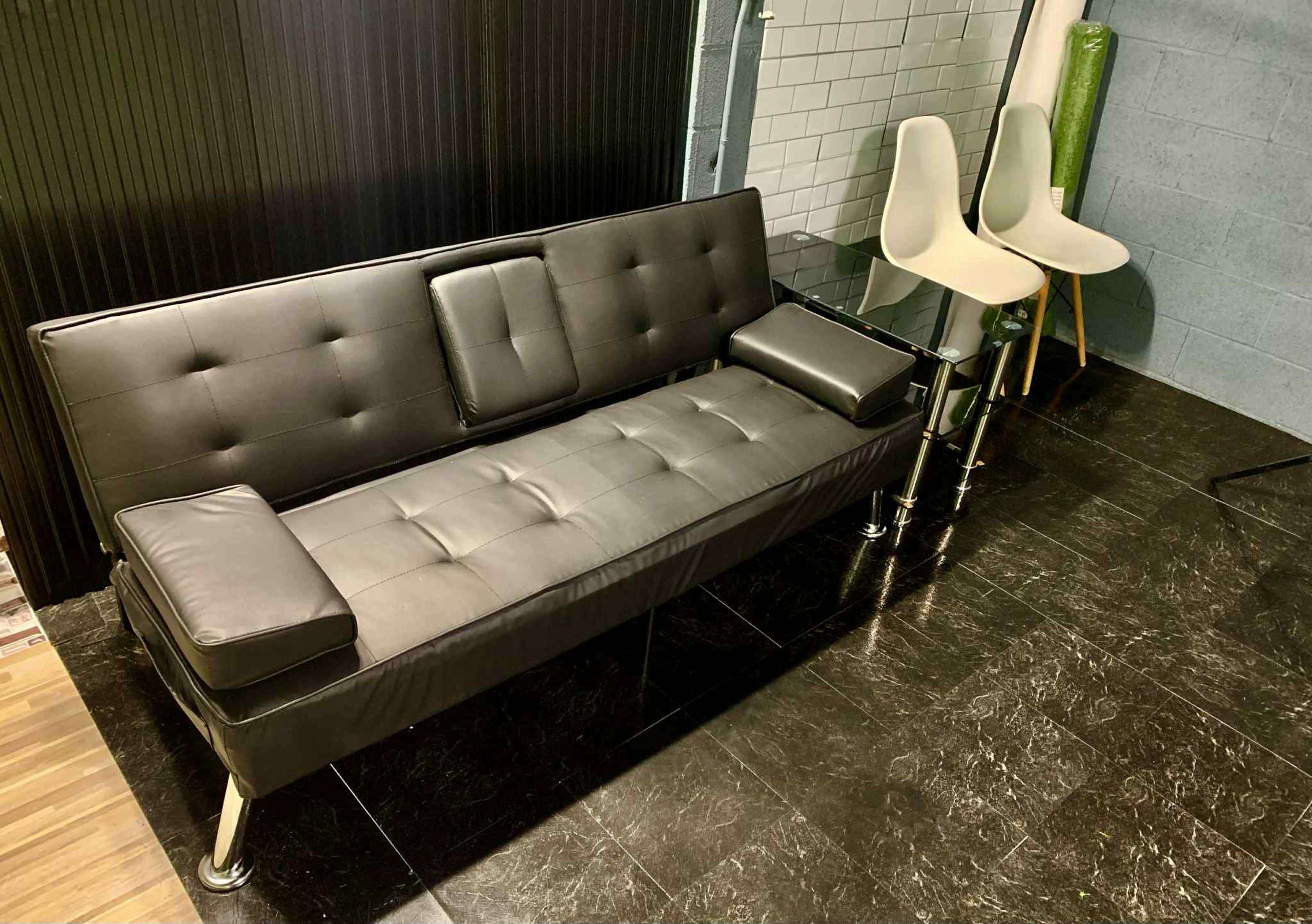 The image shows a black futon sofa with cushions, placed next to a modern glass table and two white chairs in a sleek, minimalistic setting.