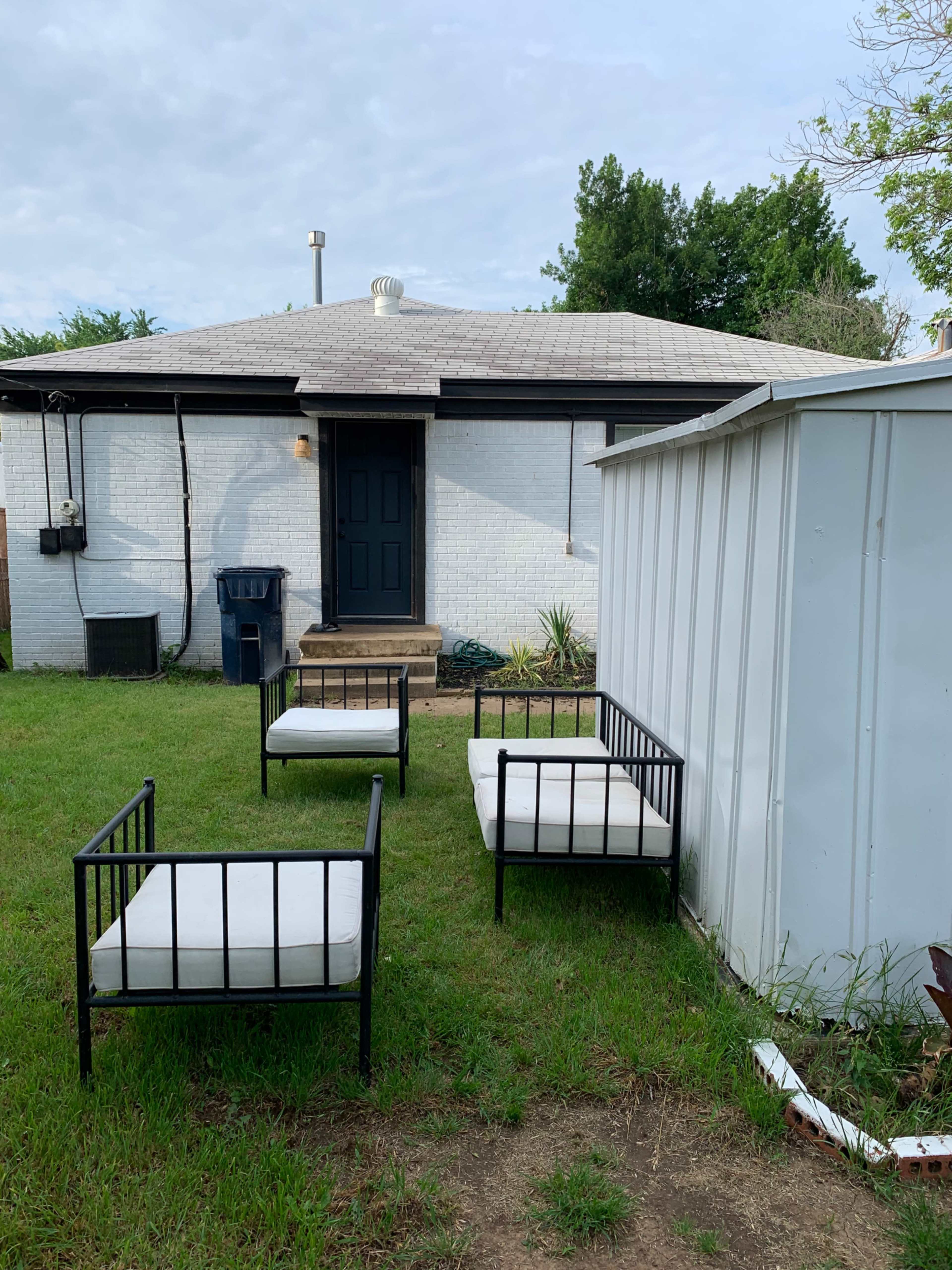 Three outdoor chairs are arranged on the grass in front of a white house with a dark door and a nearby storage shed.
