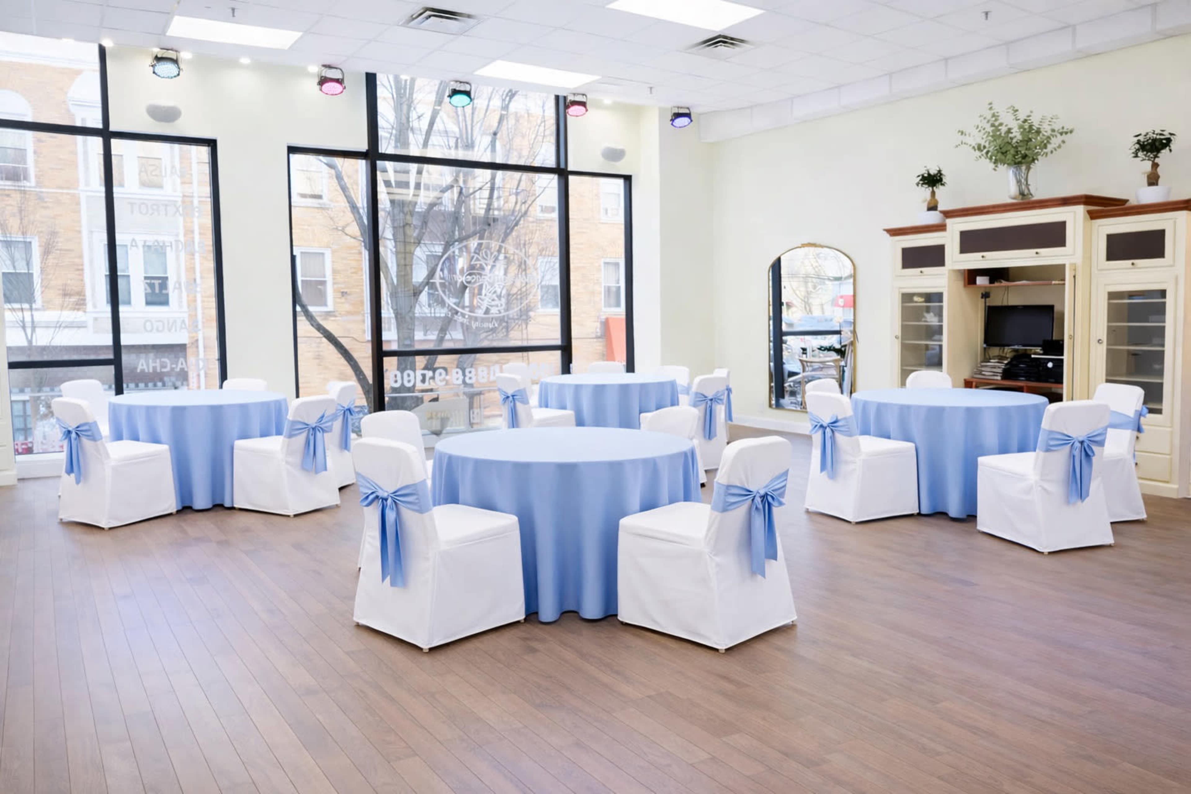 The image shows a bright event space with several round tables covered in light blue tablecloths and white chairs with blue bows, arranged around a light wooden floor.