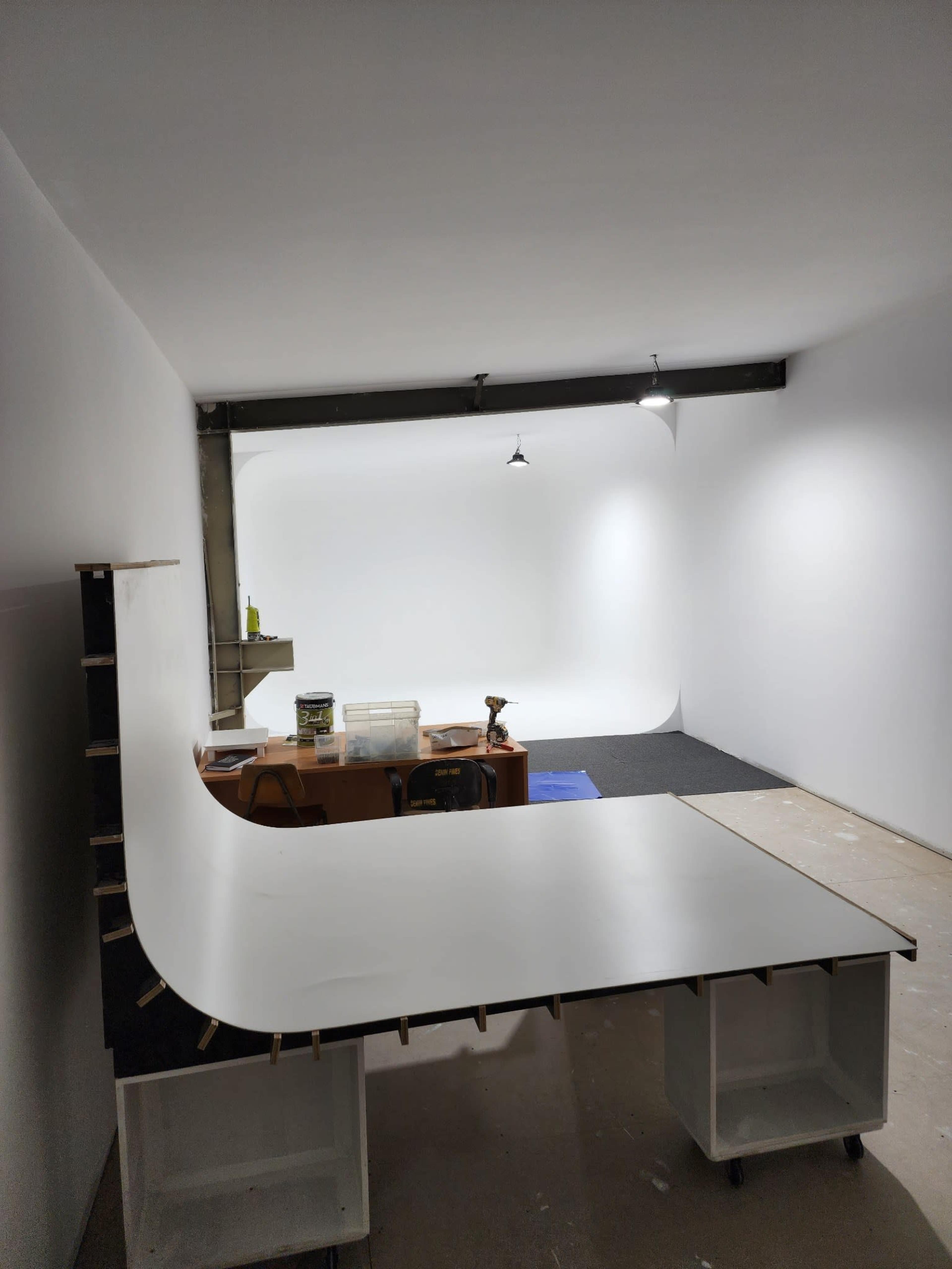 The image shows a minimalist interior of a photo studio featuring a white backdrop, a work table, and some tools.