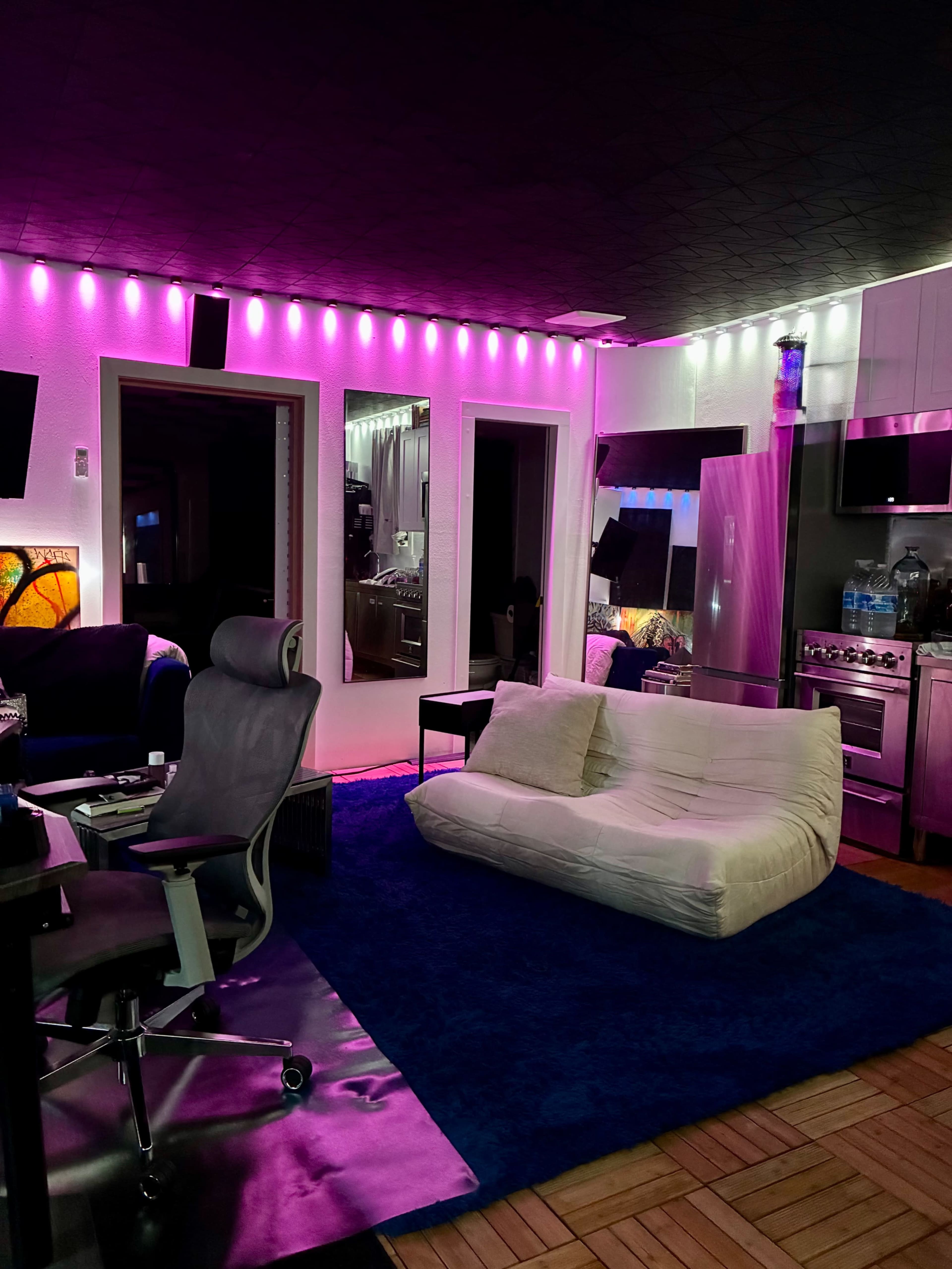 The image shows a modern room with pink ambient lighting, featuring a white couch, a blue rug, and stainless steel kitchen appliances.