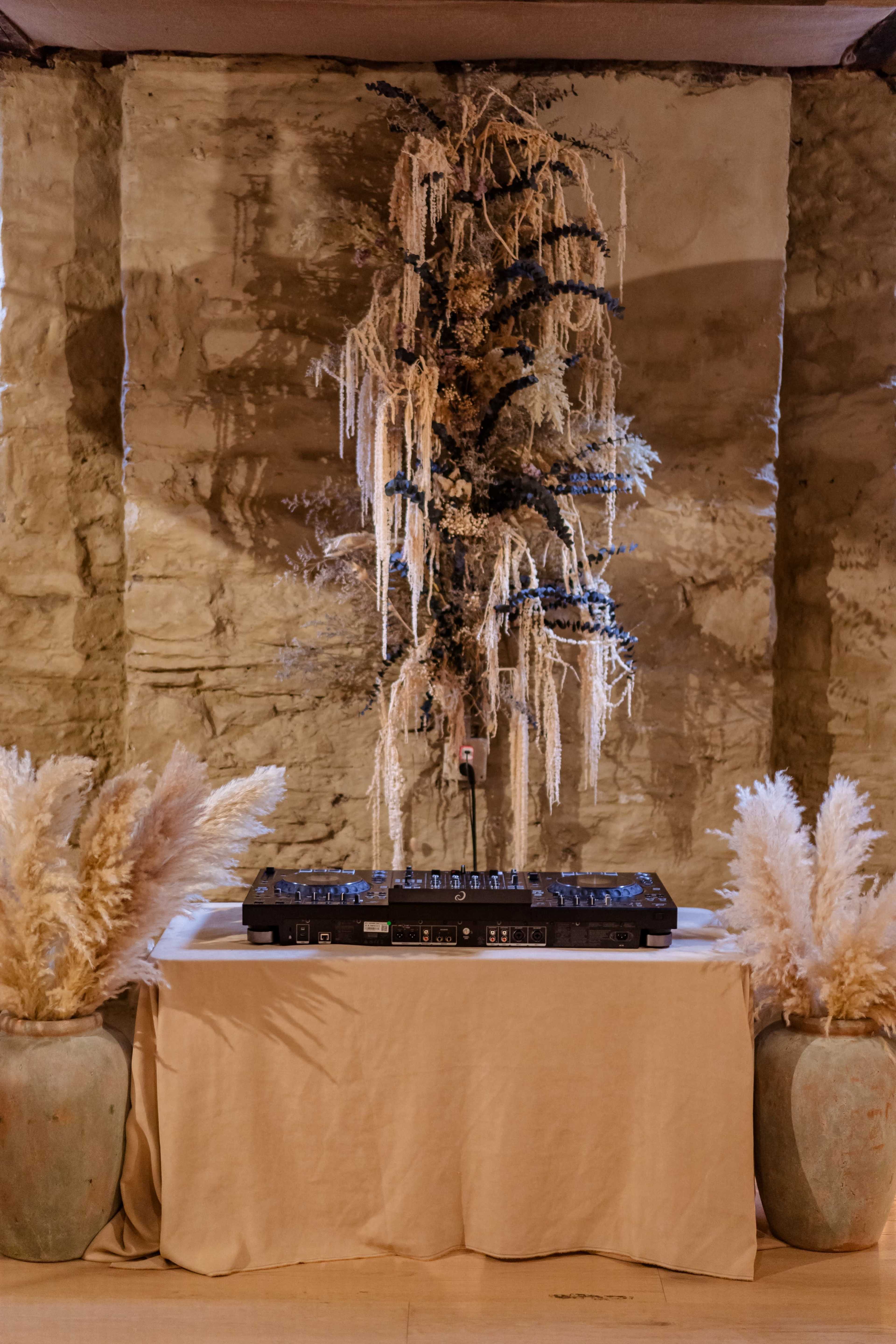 A DJ setup is placed on a table adorned with a light-colored cloth, flanked by large, decorative vases filled with dried grasses, against a stone wall backdrop.