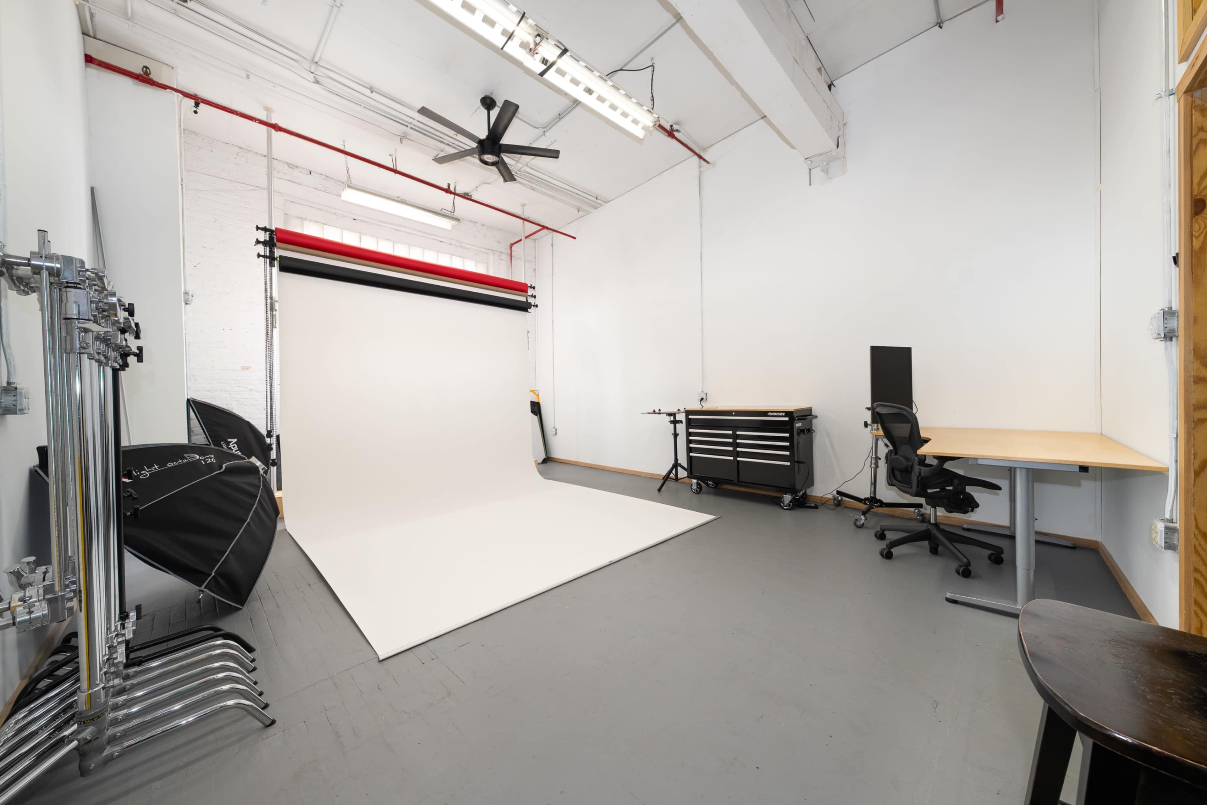 The image depicts a photography studio featuring a plain white backdrop, various lighting equipment, and a workspace with a desk and chair.