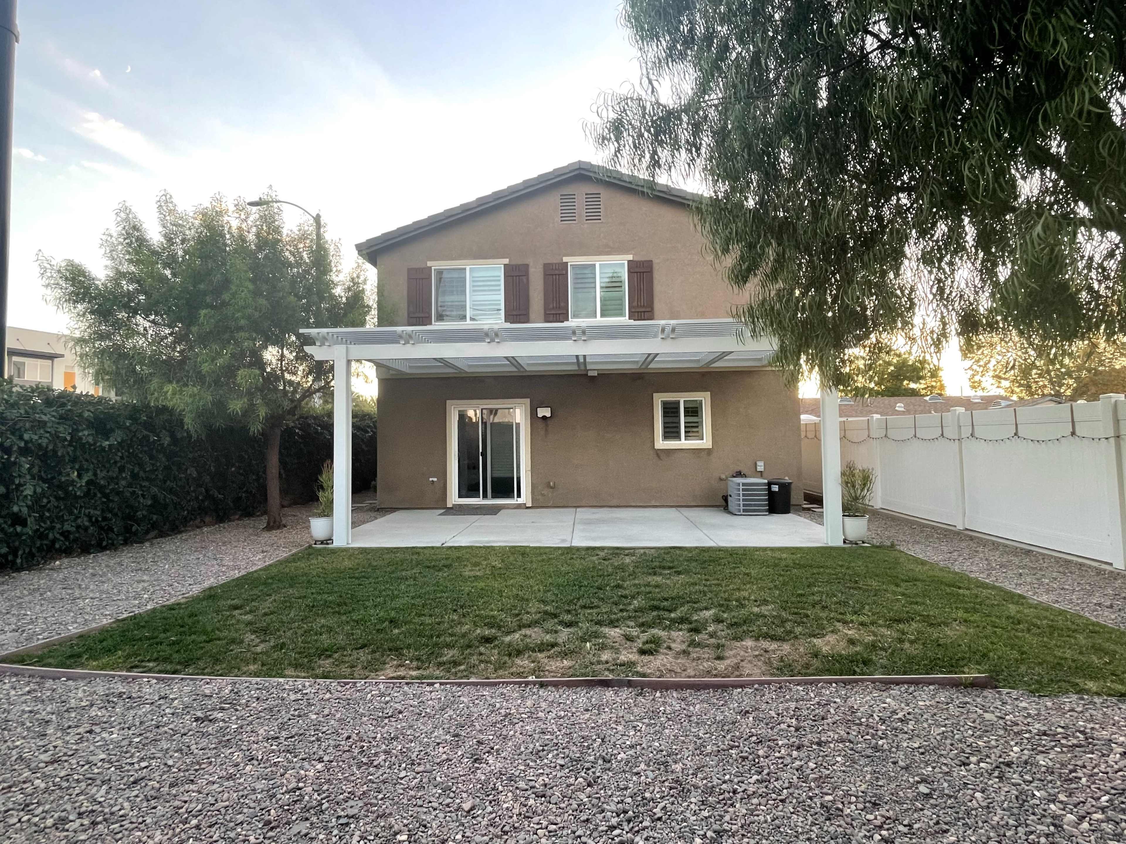 A two-story house with brown siding and shutters stands behind a grassy area, featuring a concrete patio and a white fence.