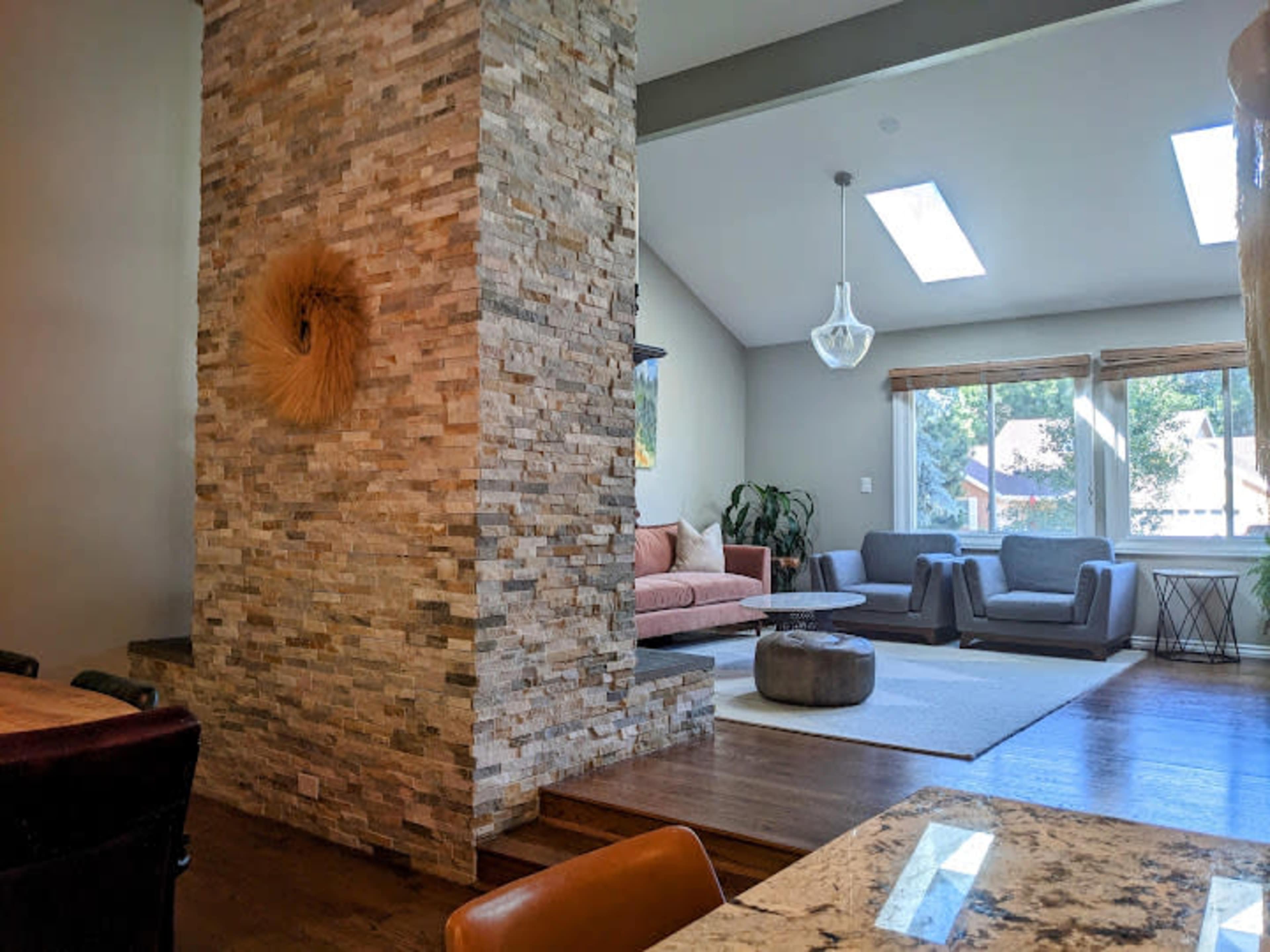 A living space divided by a stone wall, featuring a pink sofa and gray chairs illuminated by natural light from skylights.