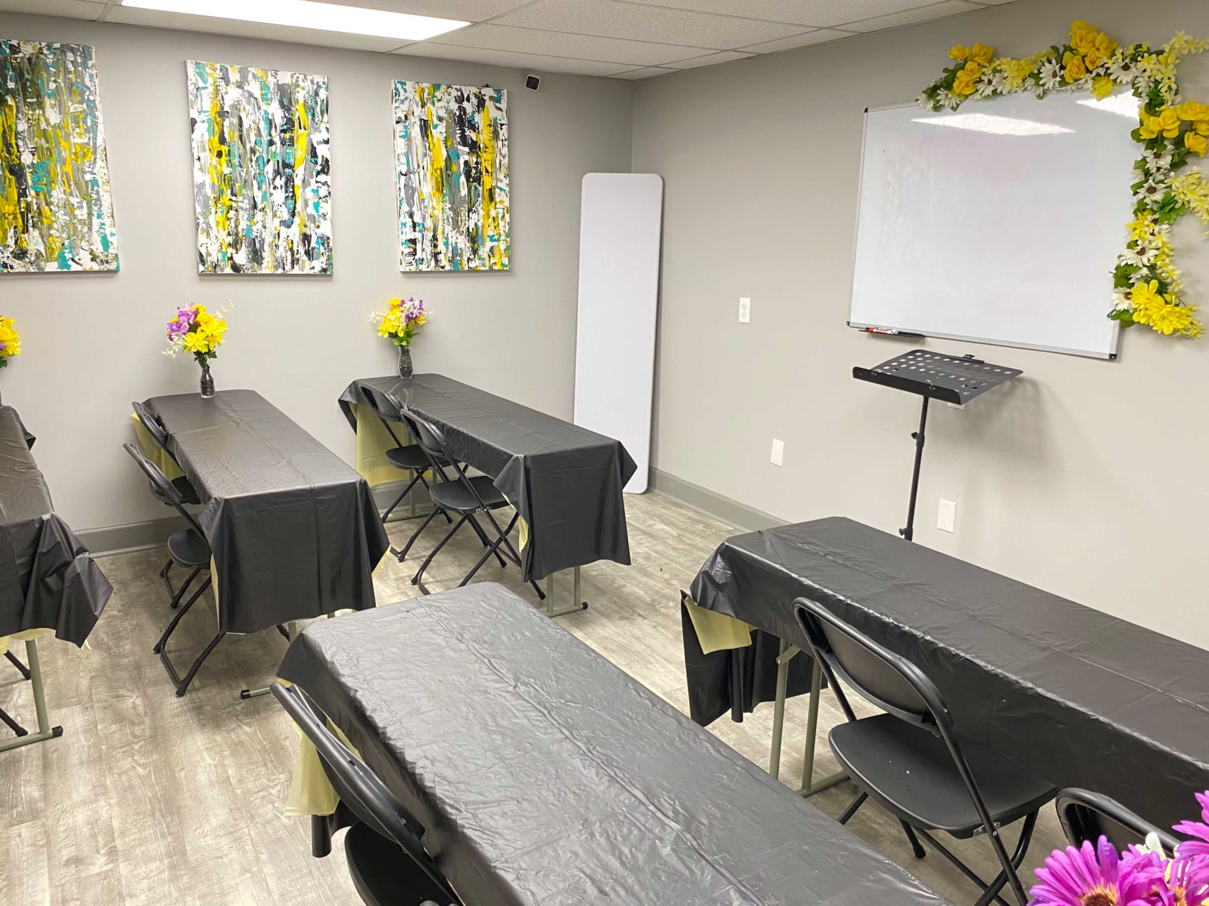 A classroom setting featuring several black rectangular tables arranged in rows, each adorned with flower centerpieces, and a whiteboard mounted on the wall alongside colorful paintings.