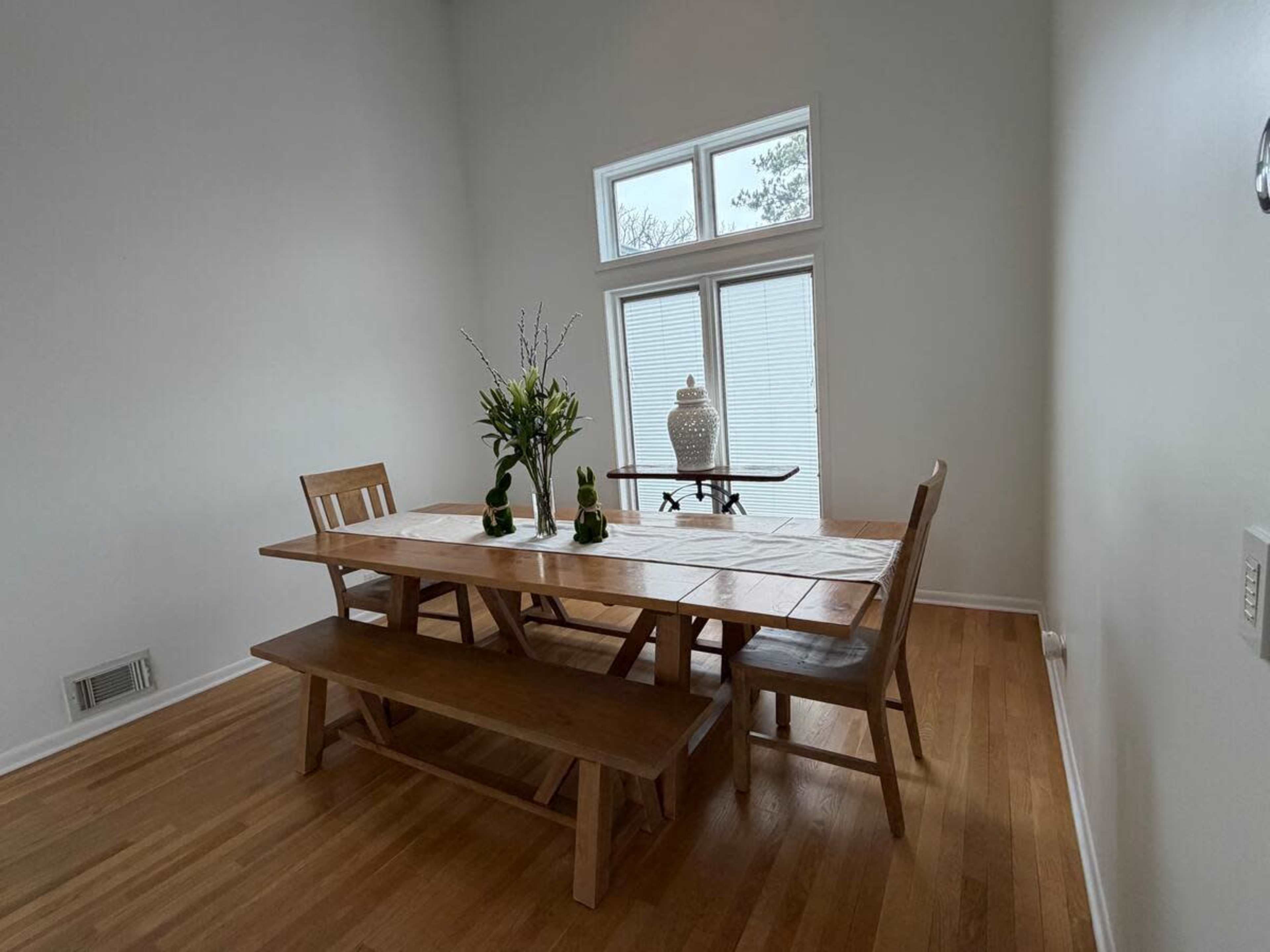 A wooden dining table with bench seating is set against a backdrop of large windows, allowing natural light to fill the room.