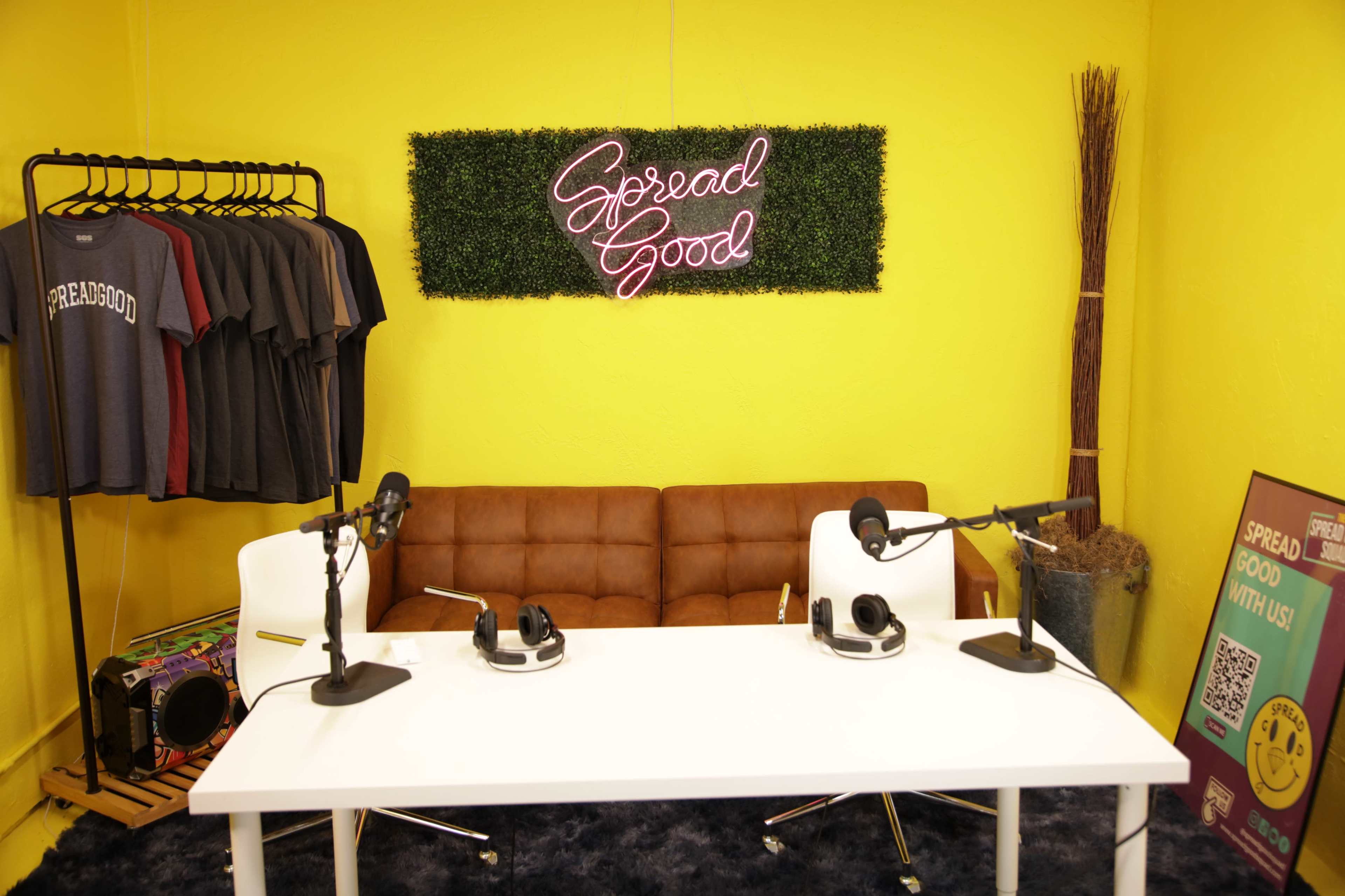 A recording studio setup features a white table with two microphones and headphones, a brown couch, a clothing rack with t-shirts, and a neon sign on a yellow wall that reads "Spread Good."