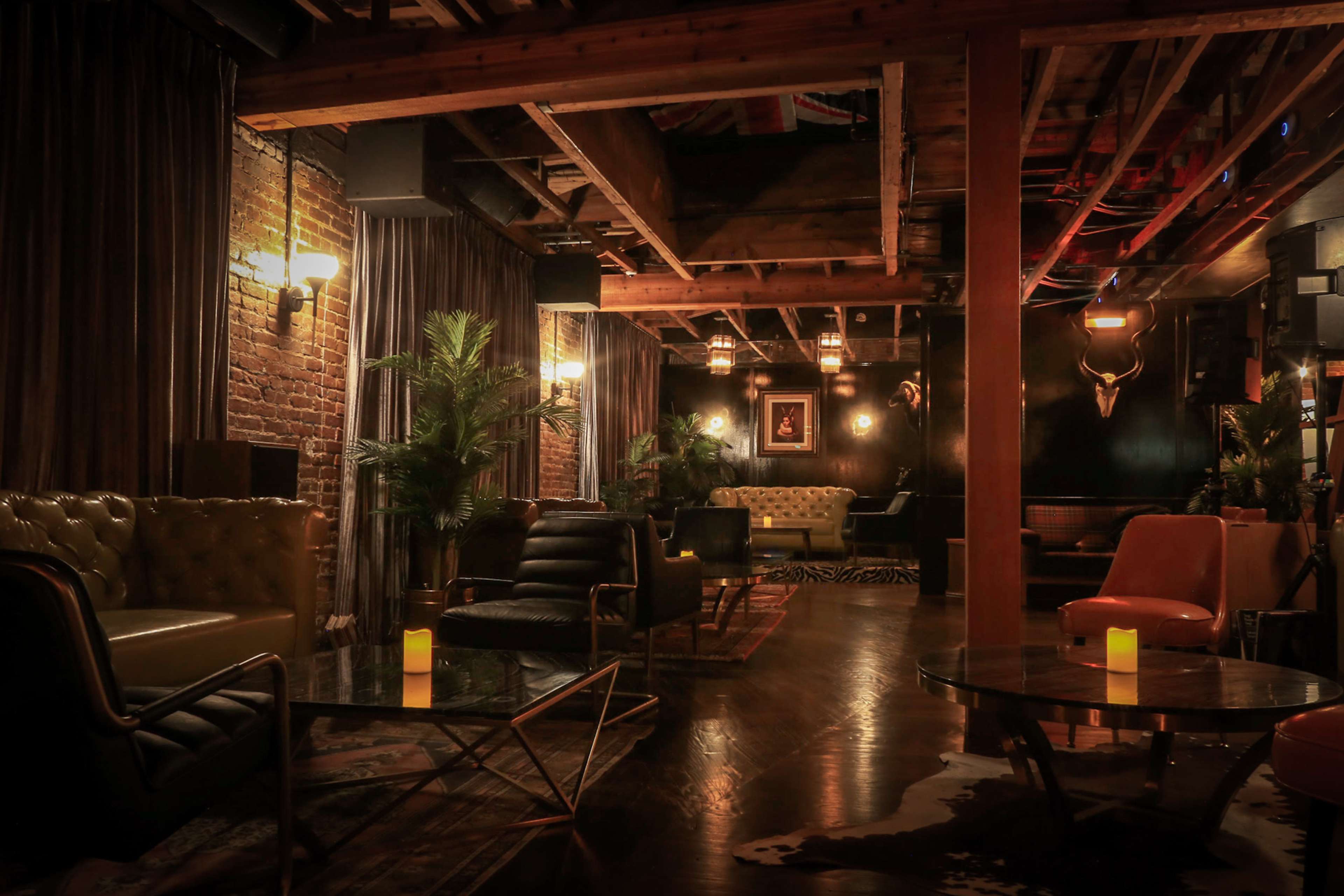 A dimly lit lounge featuring leather seating, wooden beams, brick walls, and potted plants, with candles on low tables.