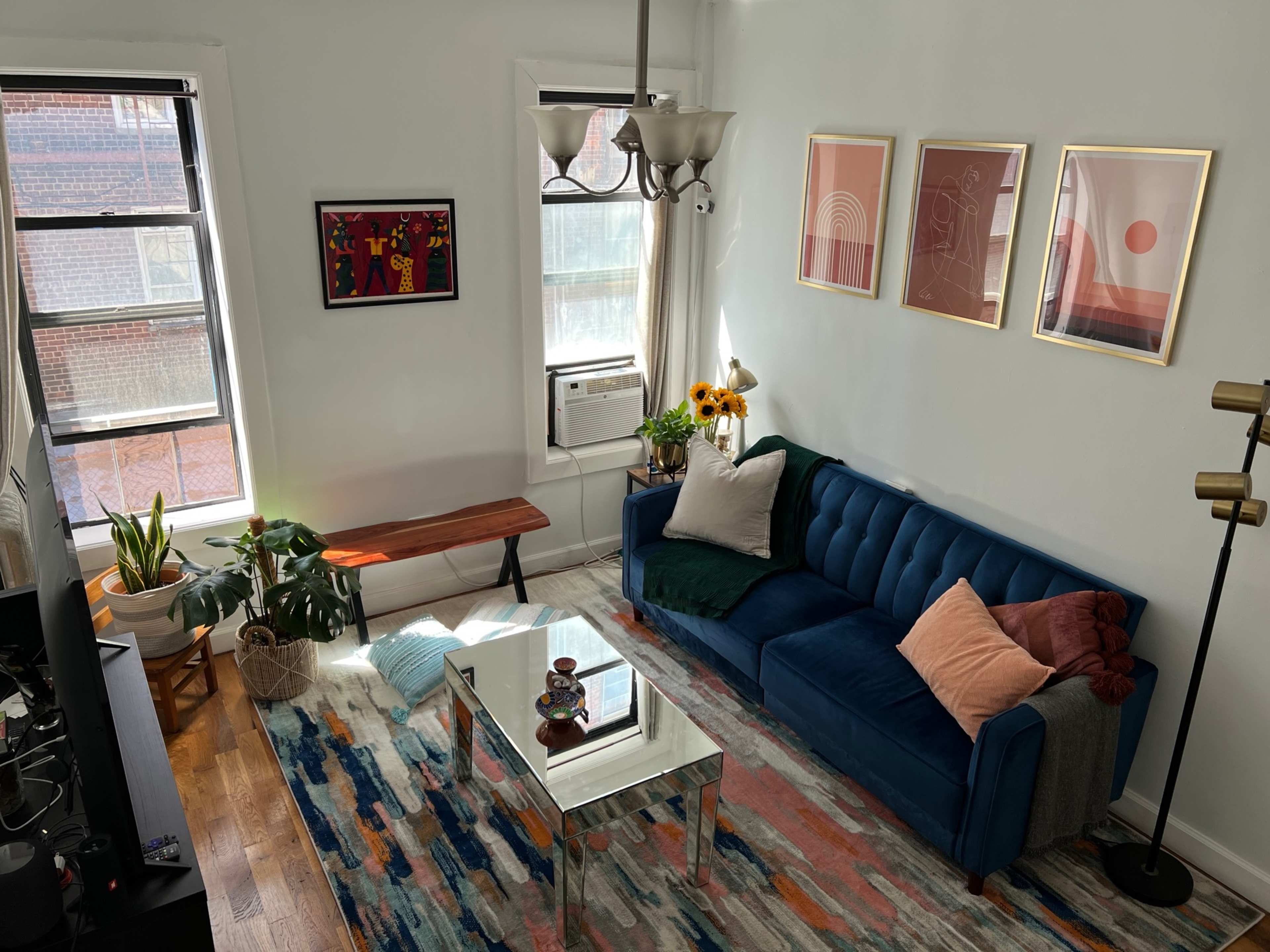 A bright living room featuring a blue couch, a wooden bench, indoor plants, and artwork on the walls.