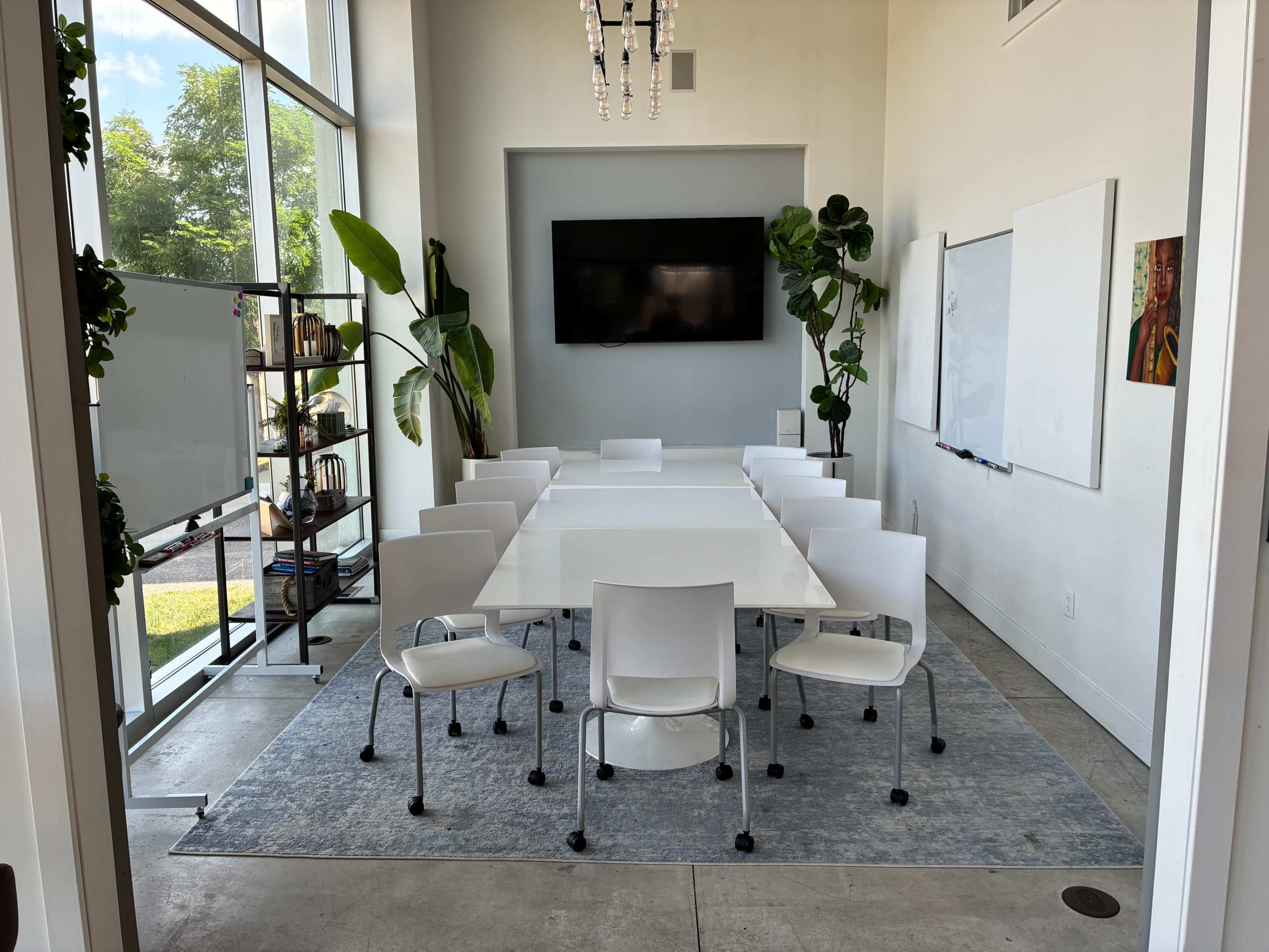 The image shows a modern conference room with a long white table surrounded by twelve chairs, large windows letting in natural light, and potted plants in the corners.