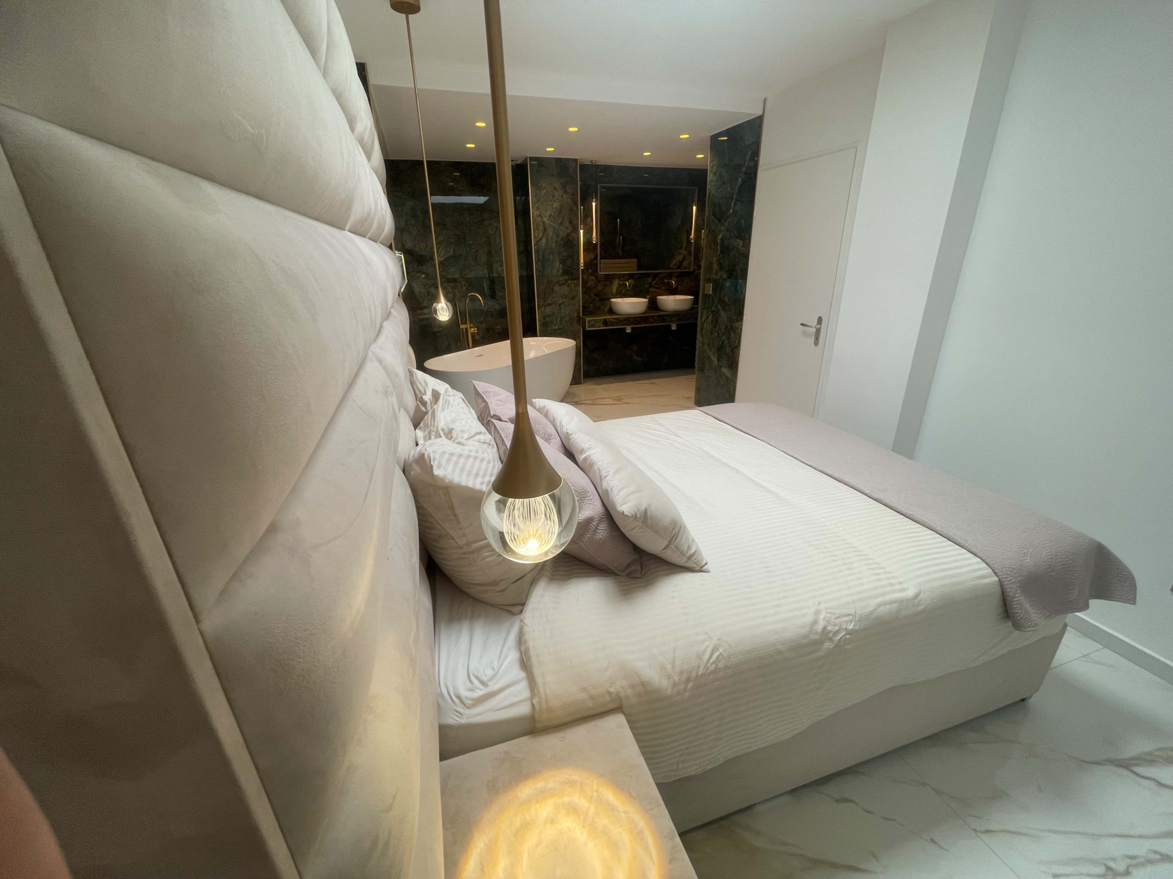 The image shows a modern bedroom with a bed featuring a tufted headboard, a bedside table with an illuminated lamp, and an adjoining bathroom visible through a glass wall.