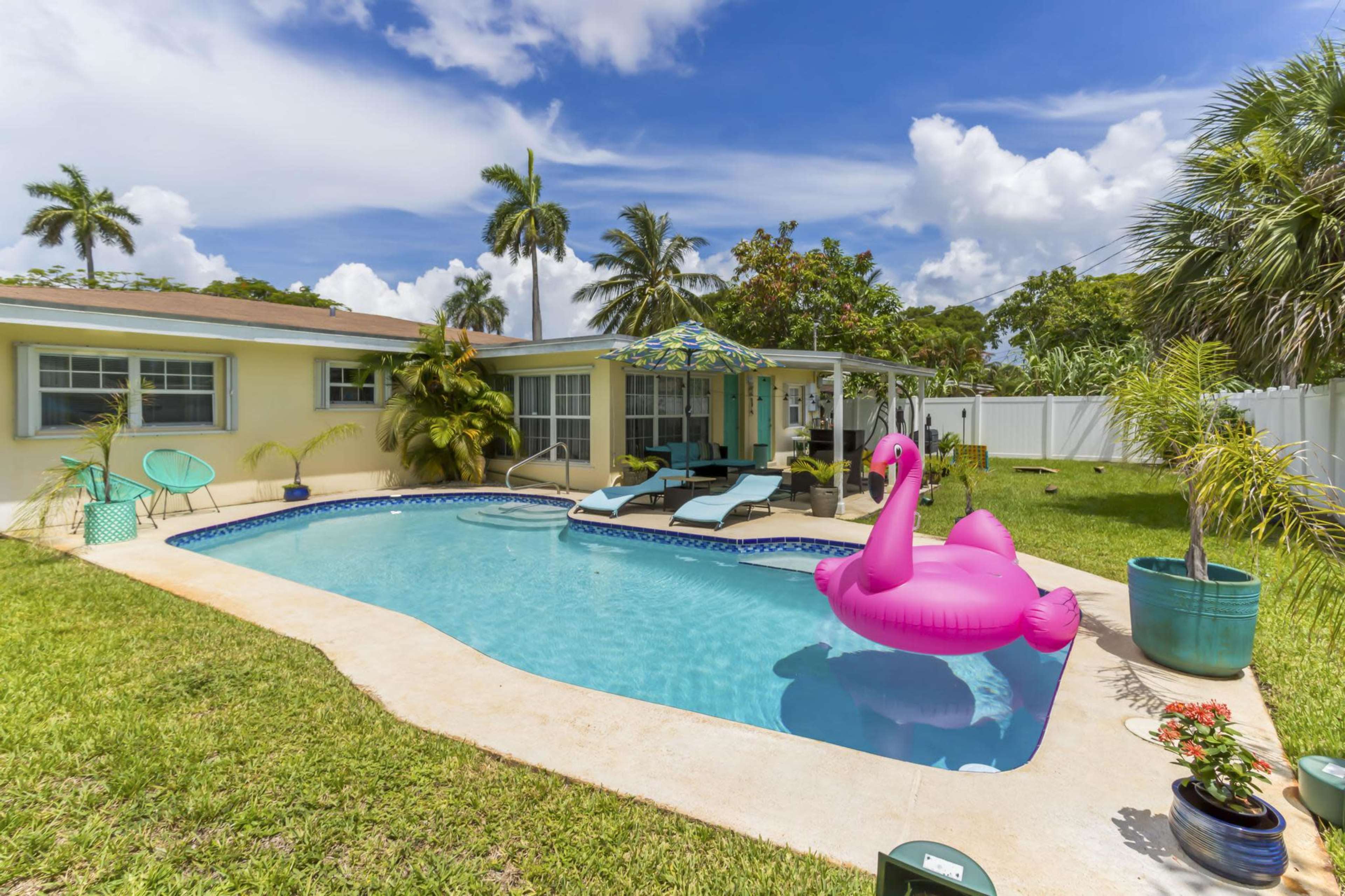 A pink flamingo floatie sits in a clear swimming pool surrounded by lounge chairs, tropical plants, and a bright blue sky with white clouds.