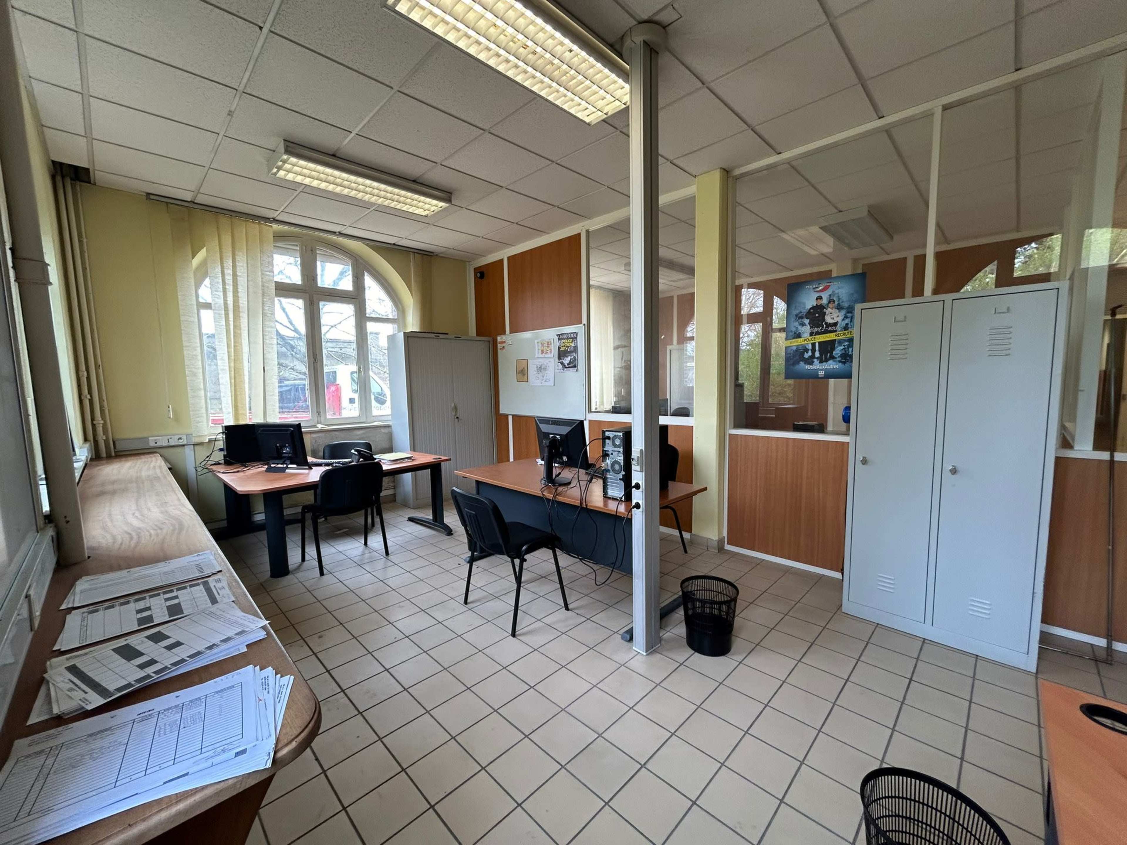 A modest office space with several desks, computers, filing cabinets, and large windows allowing natural light.