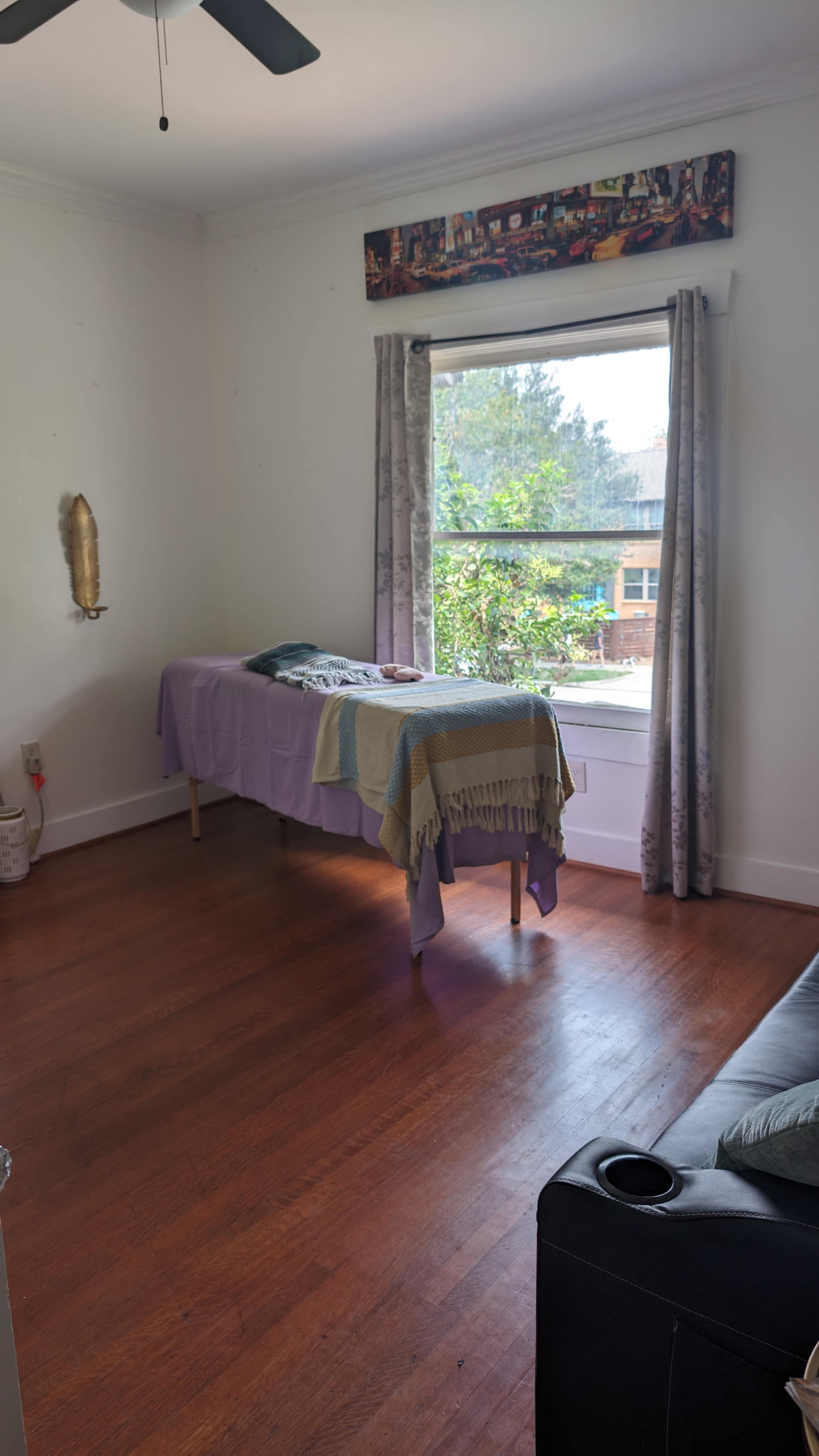 A room with wooden flooring features a table covered with a purple cloth near a window with gray curtains, and a sofa in the corner.