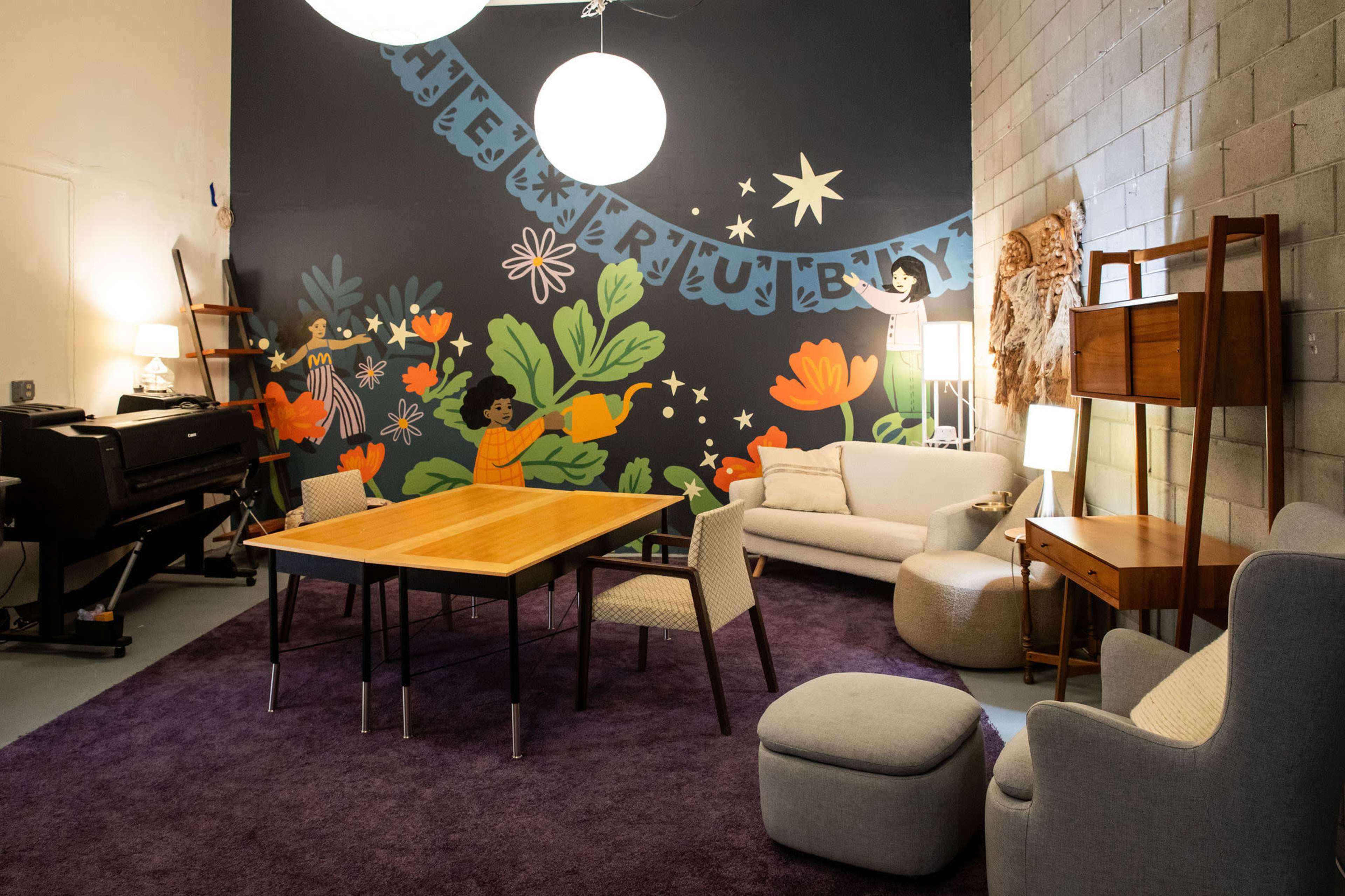 The image shows a brightly decorated room featuring a wooden table surrounded by chairs, a sofa, and various light sources, with a colorful mural on the wall.