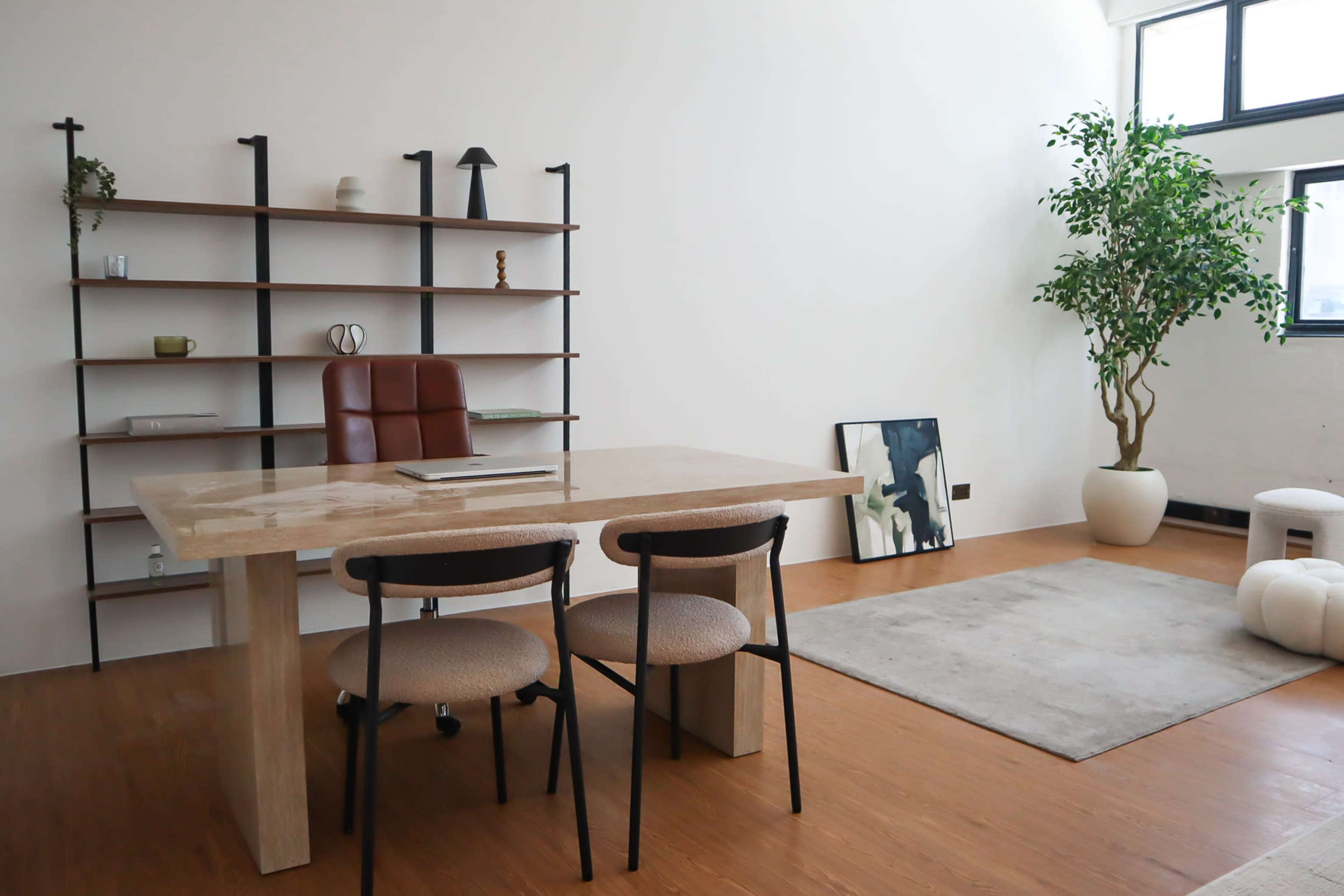 The image shows a minimalist office space featuring a large table with two chairs, a bookshelf, a potted plant, and a rug on the wooden floor.