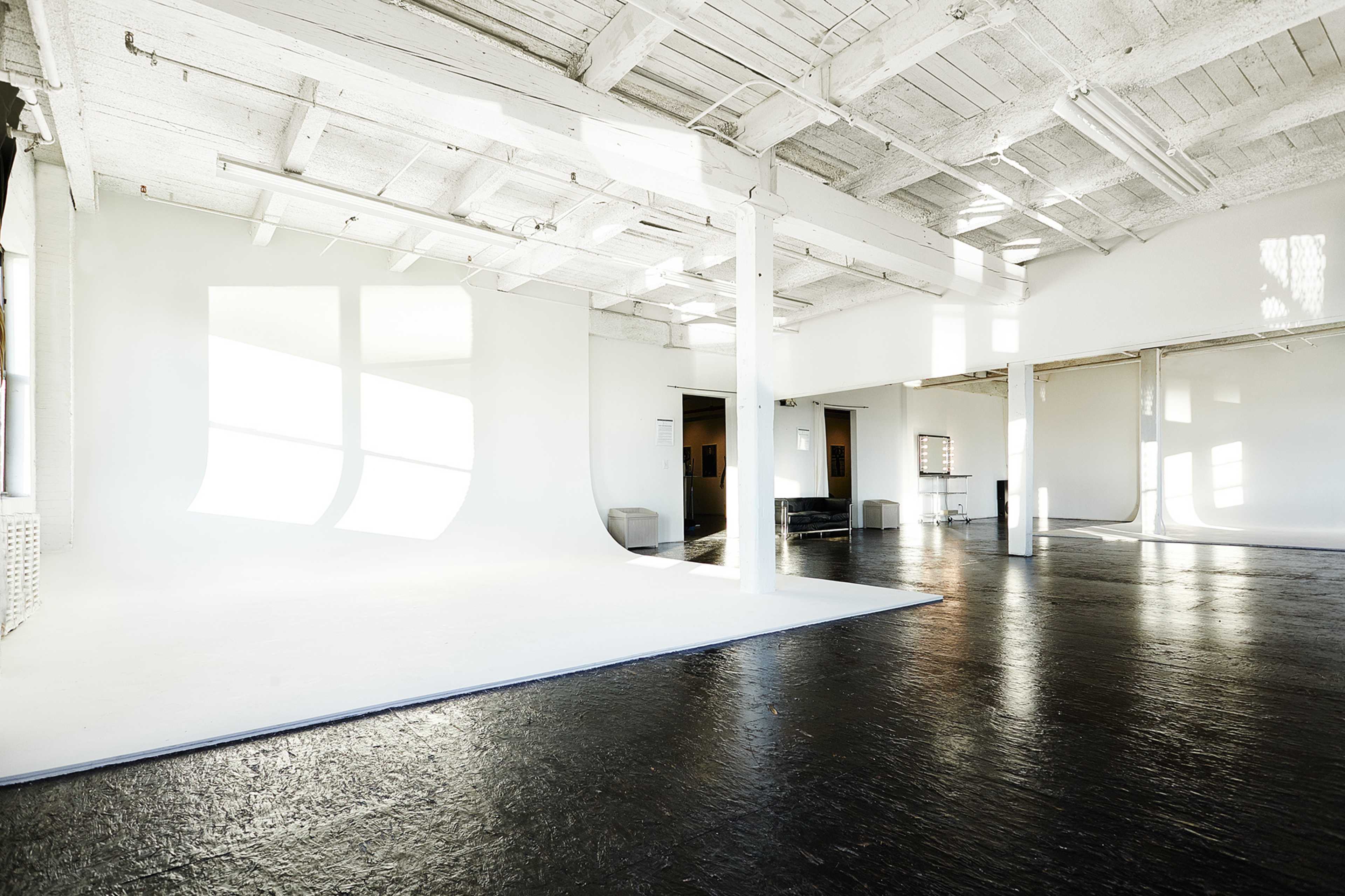 A spacious, well-lit studio with white walls, a light-colored floor, and large windows casting shadows on the surfaces.