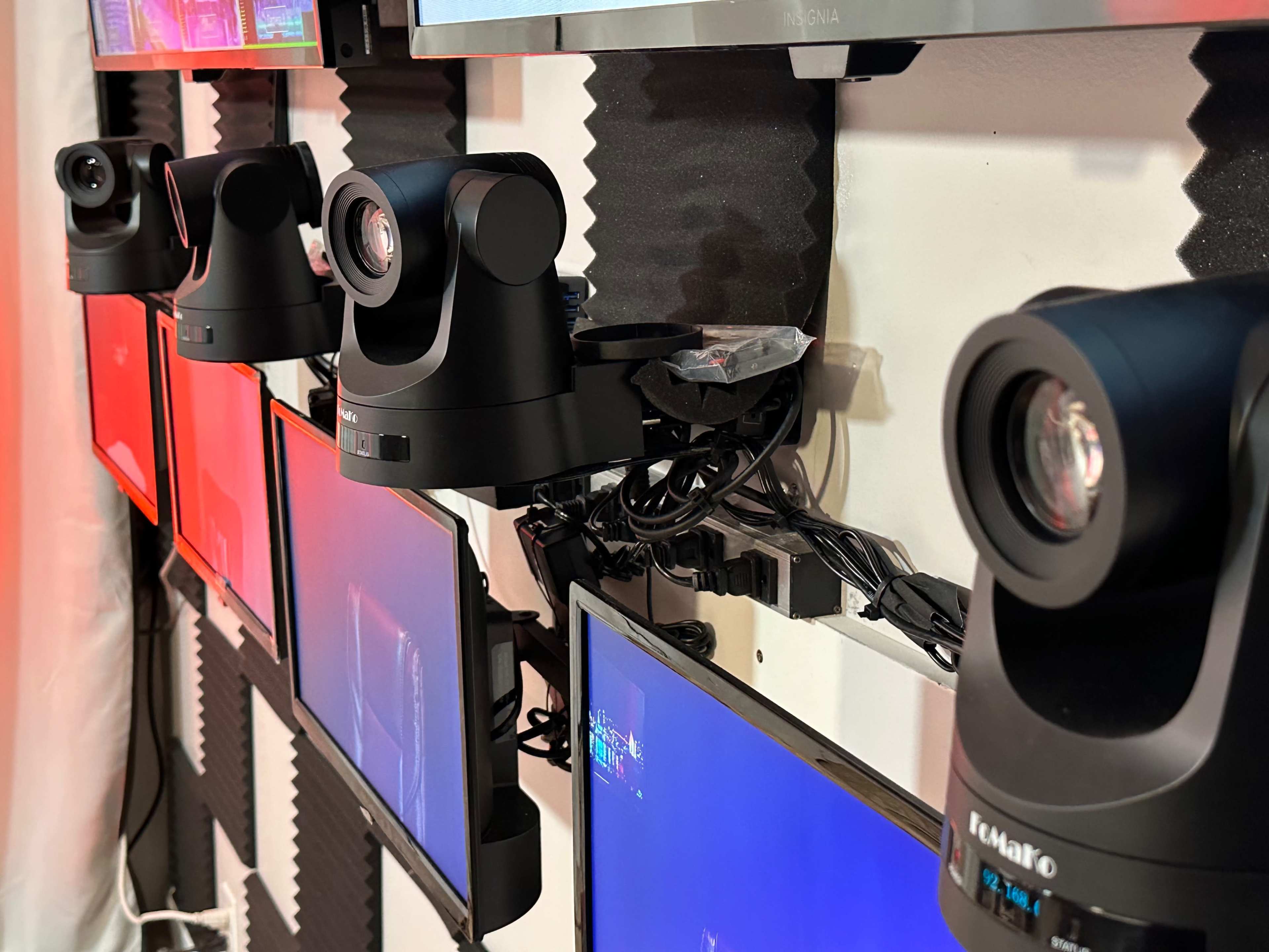 Multiple video cameras mounted on a wall above several monitors, with various colored screens illuminated.