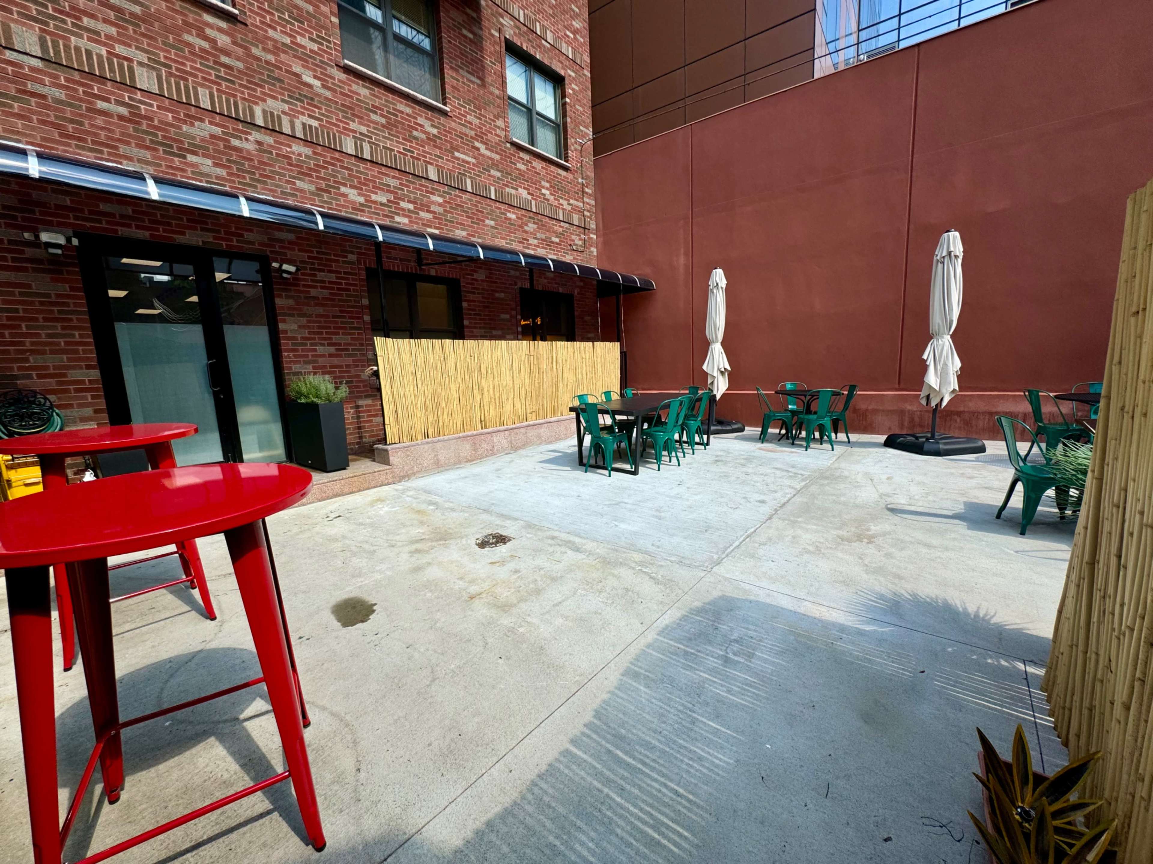 A sparsely furnished outdoor patio area with green tables and chairs, two umbrellas, and a bamboo fence against a brick building.