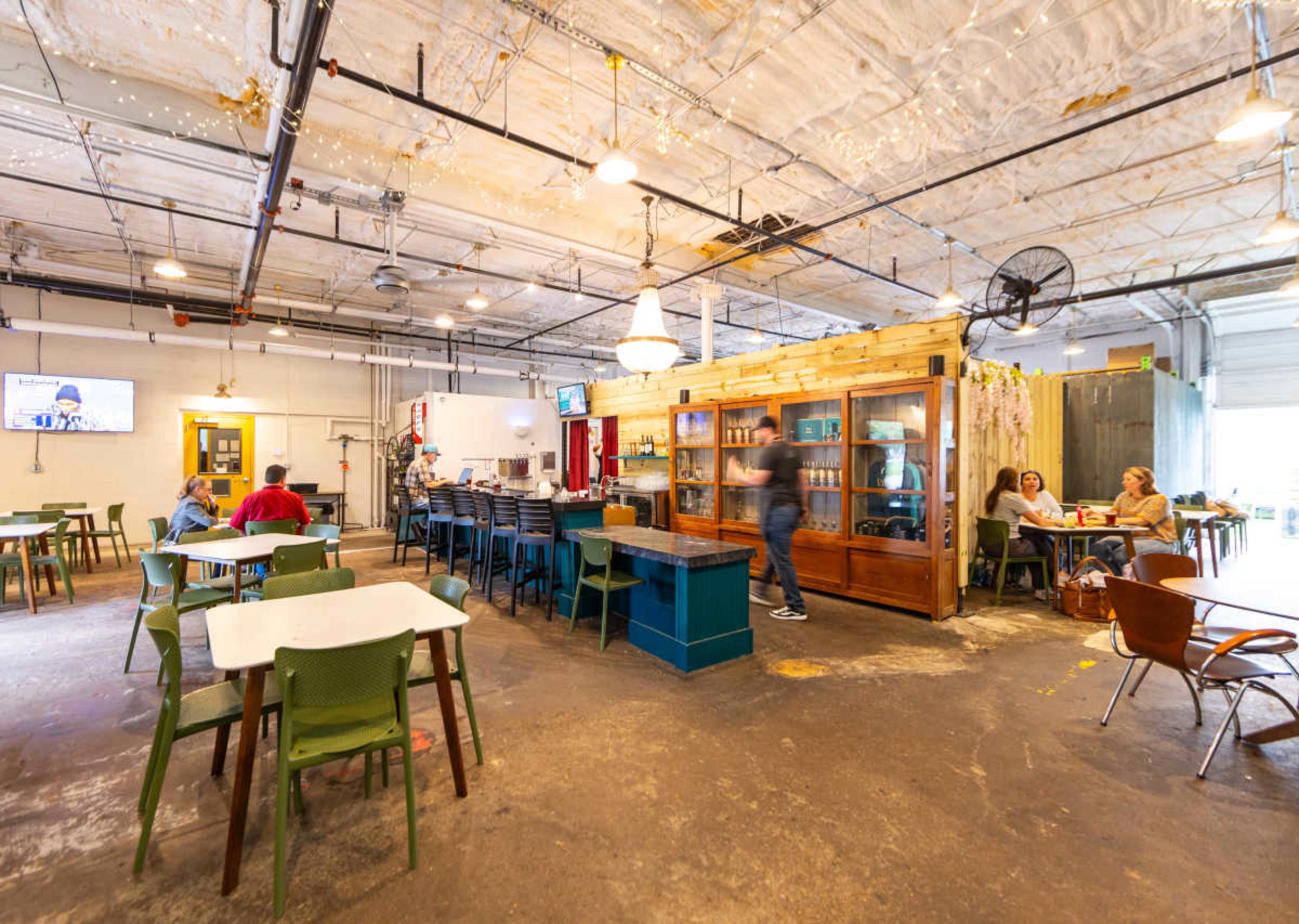 The image shows an indoor café space with tables and chairs, a service area with a counter, and patrons sitting and interacting.