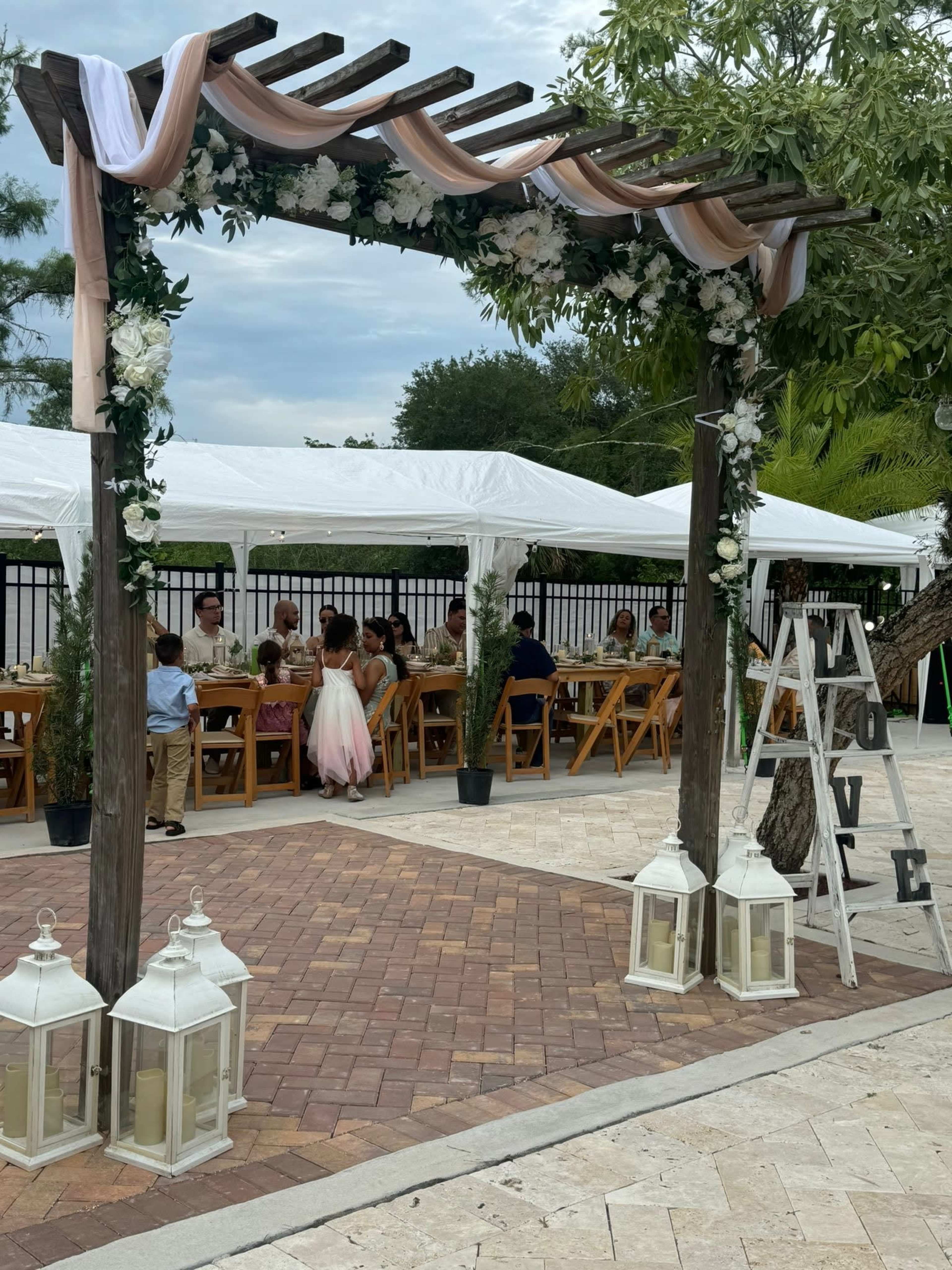 A decorated wooden pergola adorned with flowers and drapes stands in front of a tented area where guests are seated at long tables.