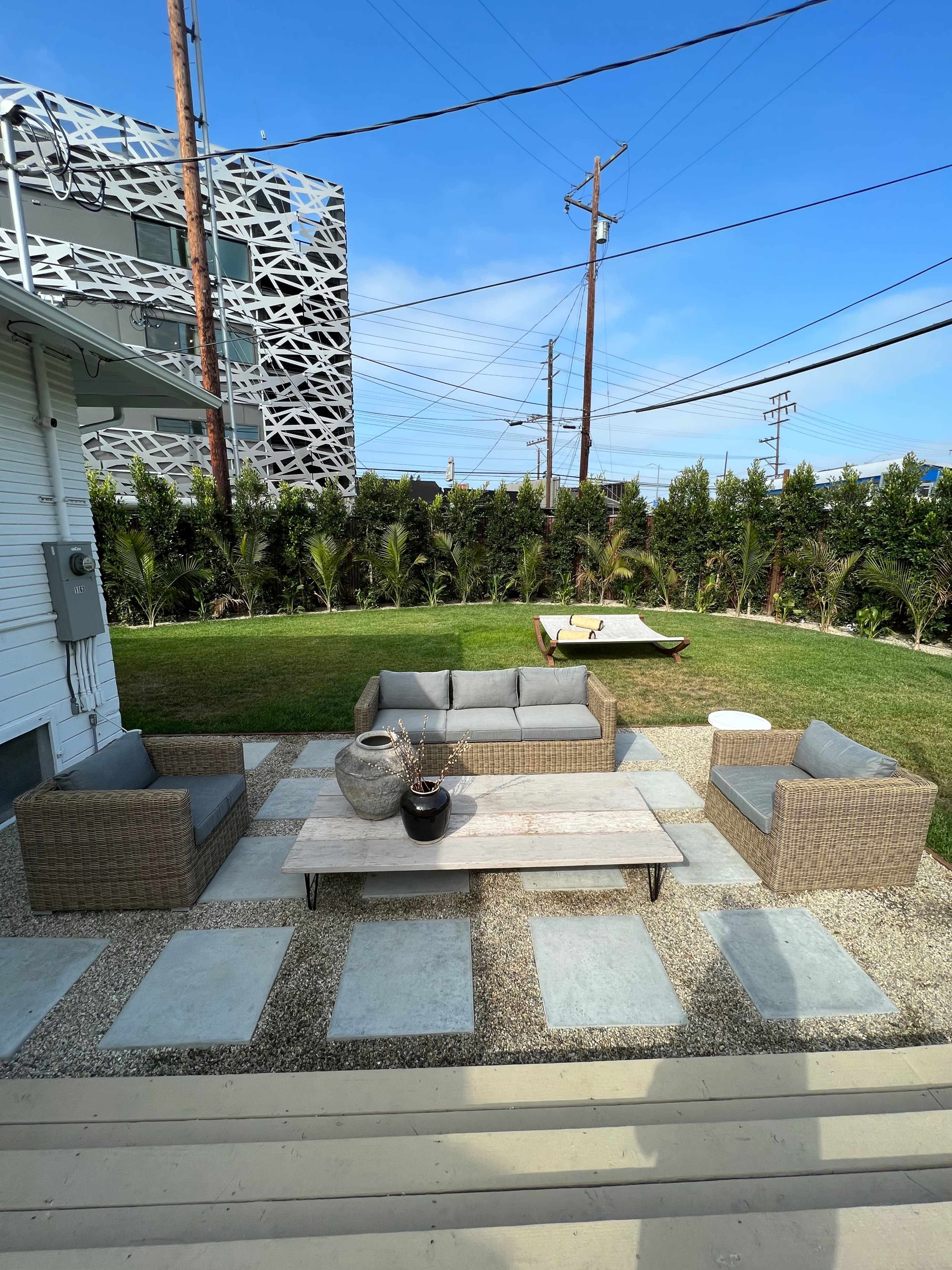 A patio area features a gray sectional sofa set and a wooden coffee table, surrounded by stone pavers and a green lawn with palm trees in the background.
