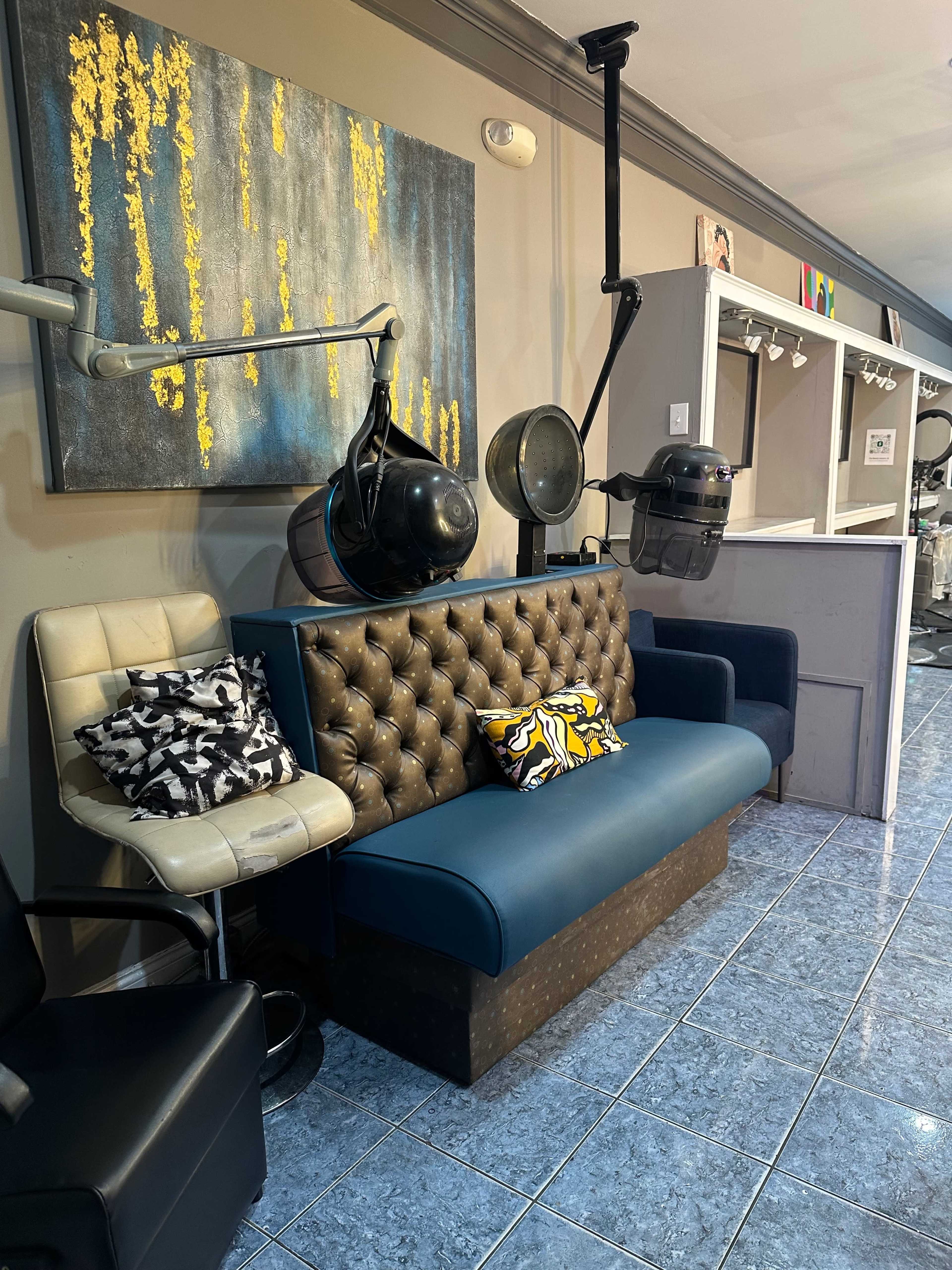 A hairstyling salon with several salon chairs and hairdryers mounted on poles beside a patterned couch and a wall art piece.