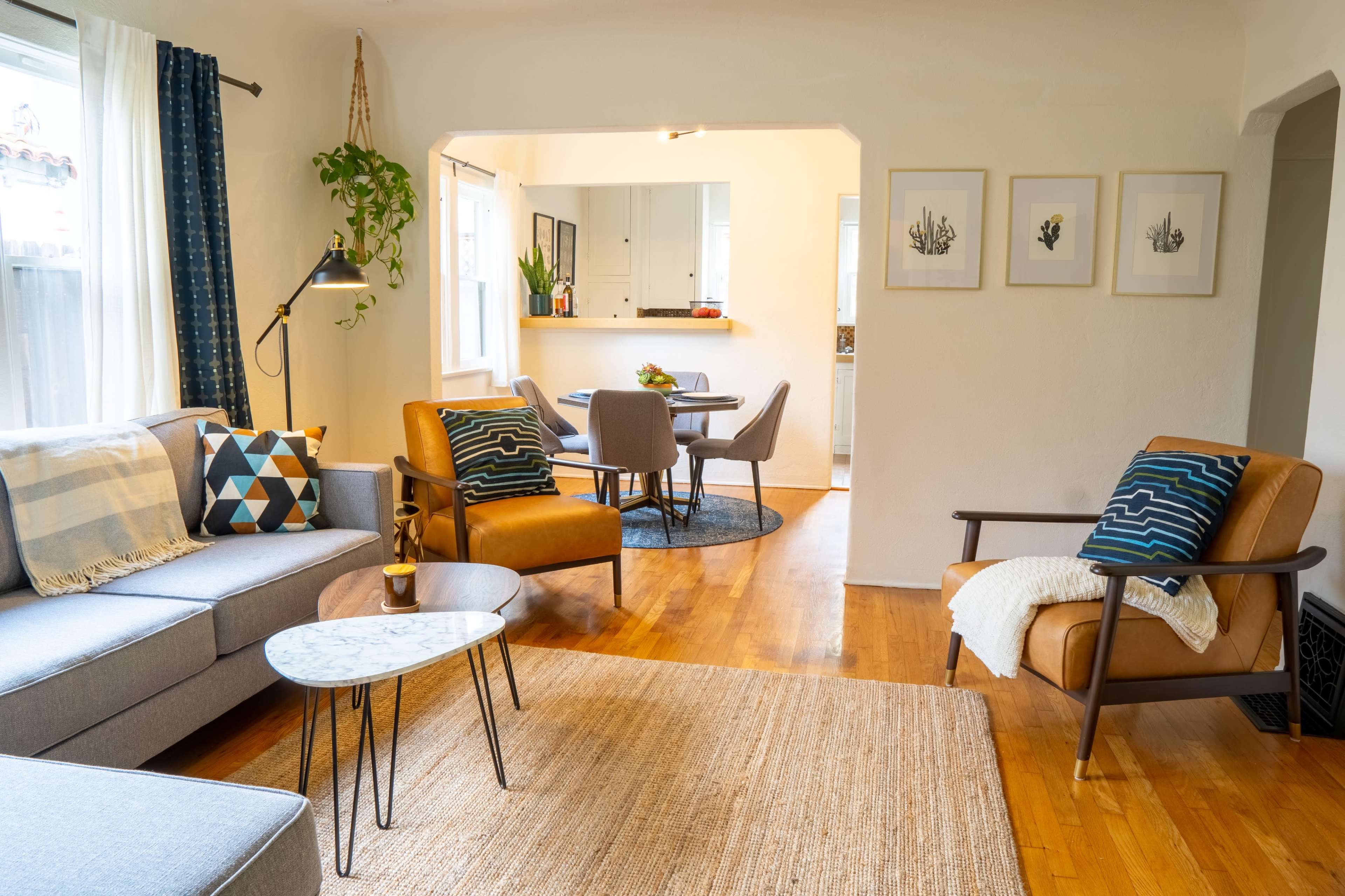 A modern living room features a gray sofa, two brown armchairs, a marble coffee table, and a jute rug, with a dining area visible in the background.