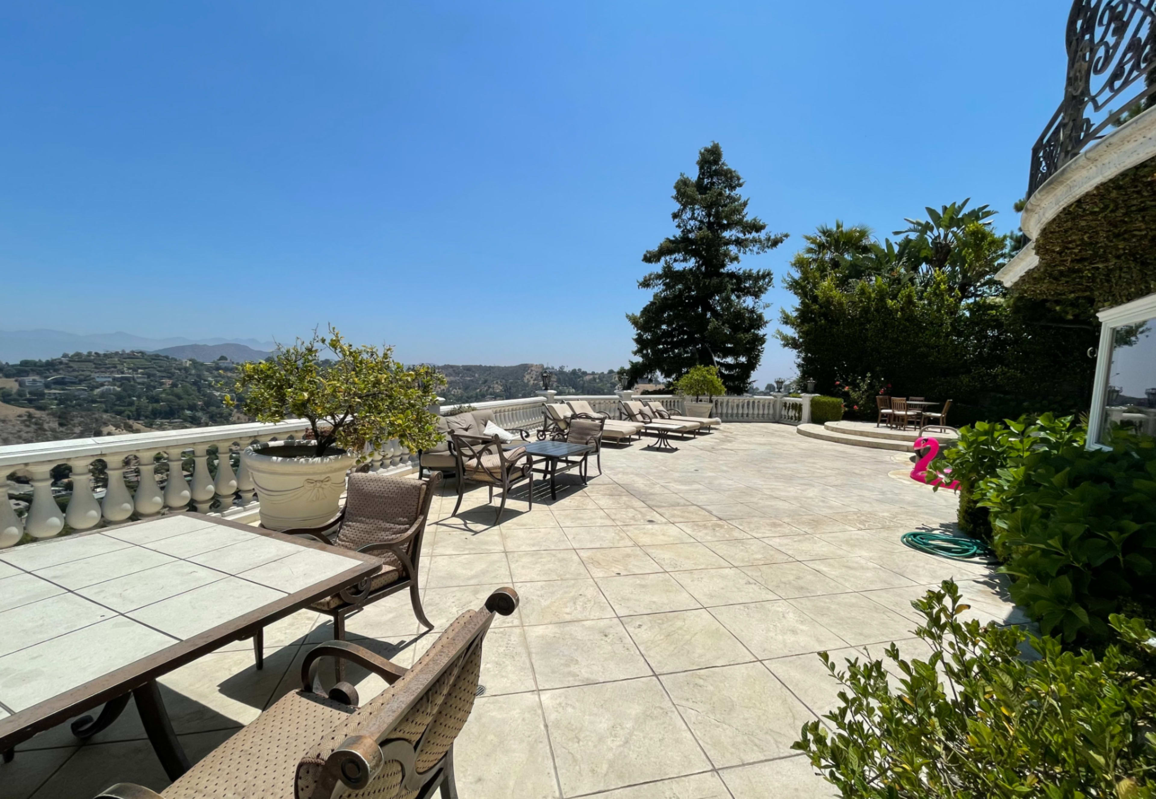 A large outdoor terrace with several lounge chairs and tables, surrounded by greenery and hills in the background under a clear blue sky.